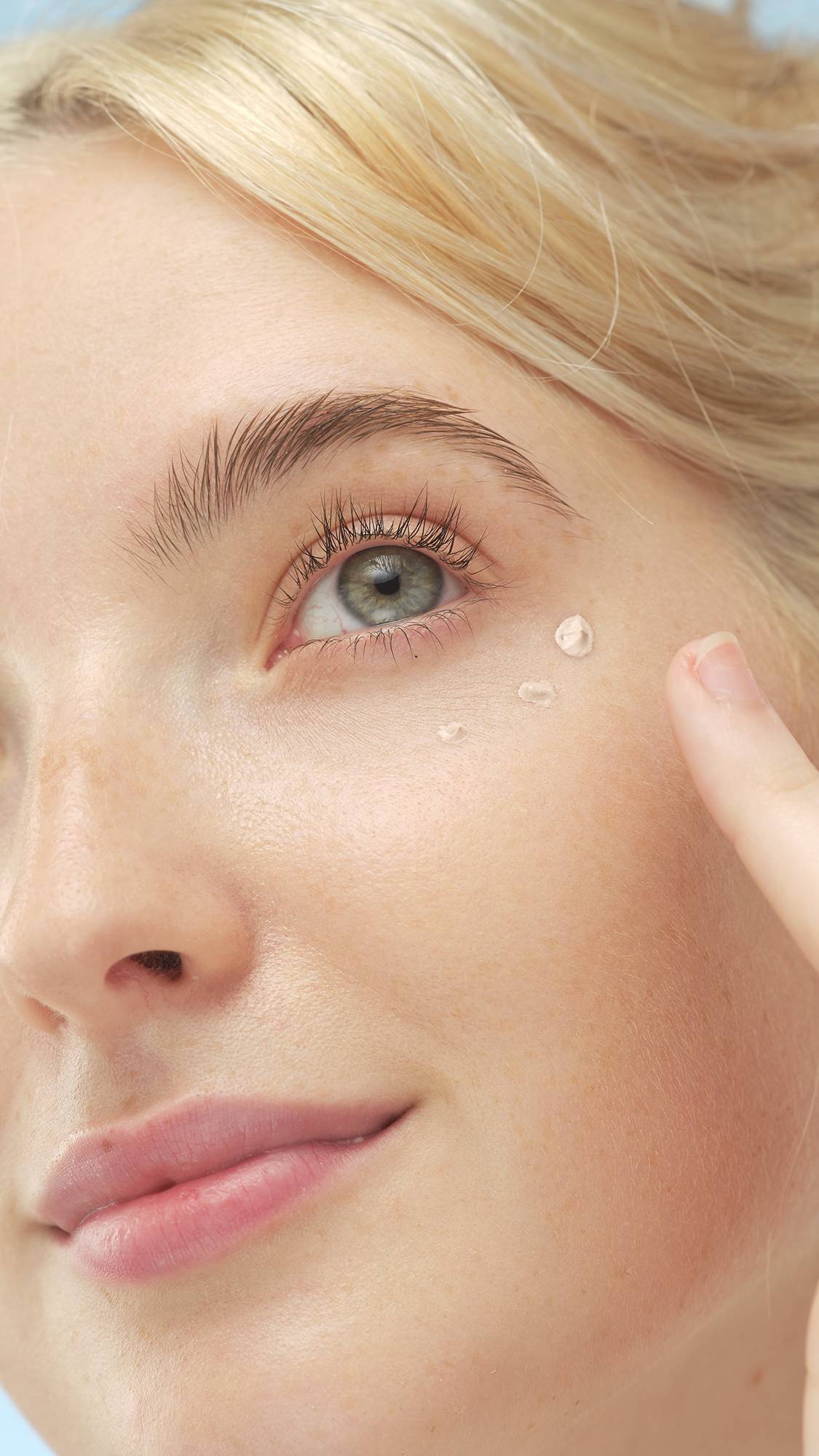 A close-up vertical shot of a person with fair skin, looking up and right. Their finger is next to three drops of Luminator - skin tint on their cheekbone, ready to be blended, highlighting their light eye and blonde hair.