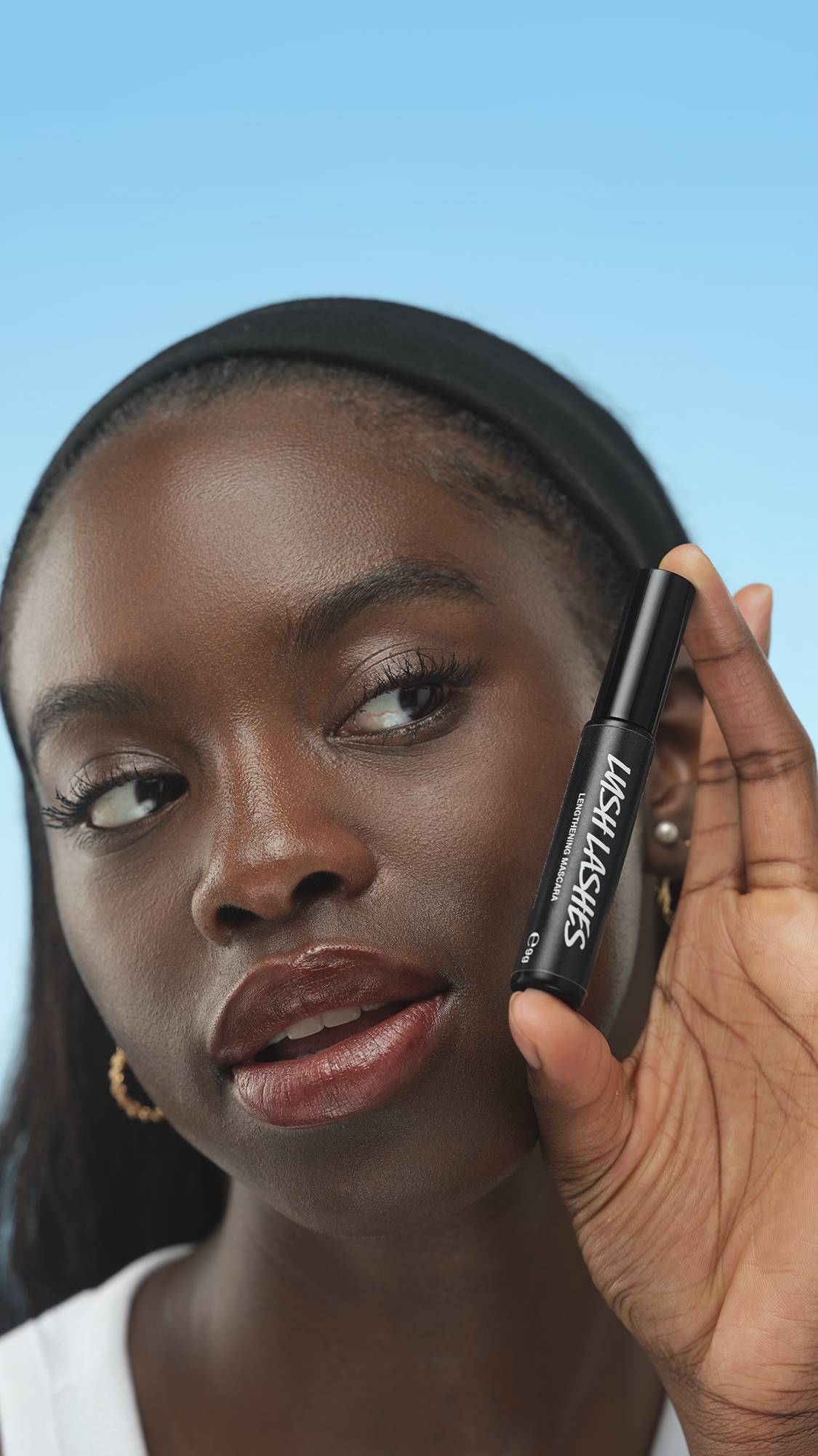 A person with dark skin and a black headband holds up Lush Lashes ~ Mascara near their face. They look to the side, wearing glossy berry lips and defined lashes, against a blue background.