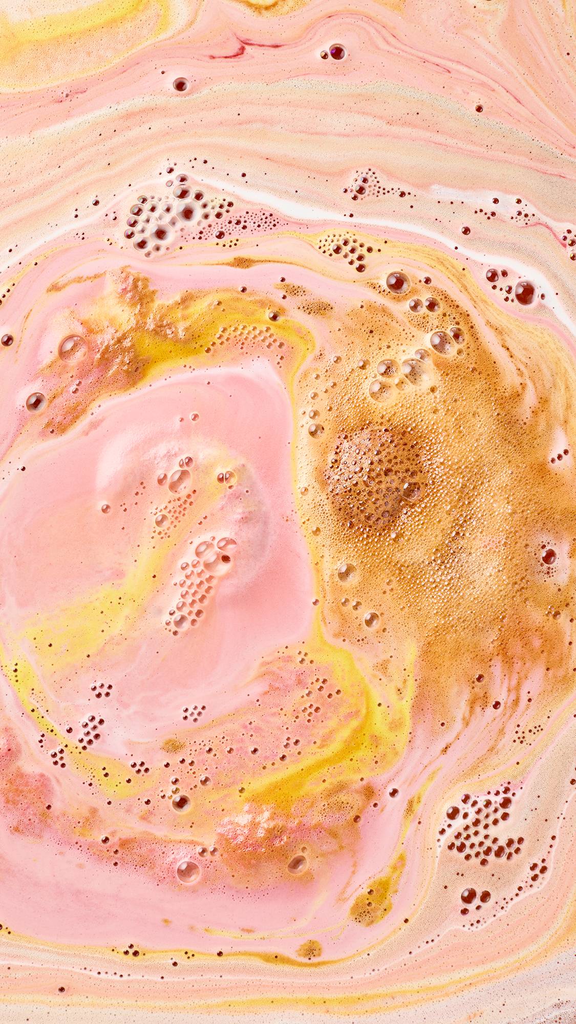 Swirling ripples of bubblegum pink, warm ochre yellow, and rich golden-brown foam merge together across silky, pastel peach bathwater as a bath bomb fully dissolves.