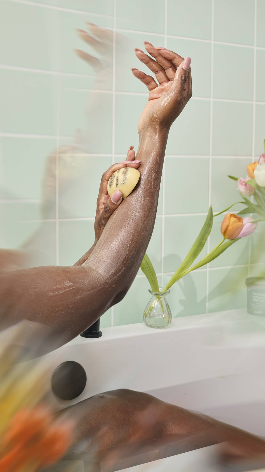 A person in a bathtub massages a "Mama-chino Scrubee" body butter bar onto their wet arm. The scene is set against mint green tiles, with a vase of orange and pink tulips resting on the edge of the white tub.