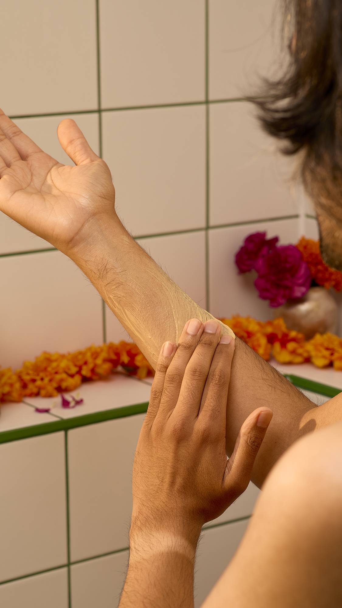A person rubs smooth, yellow Lush "Mango Kulfi" body lotion into their forearm in a tiled bathroom setting, with colourful marigold and rose flowers blurred in the background.
