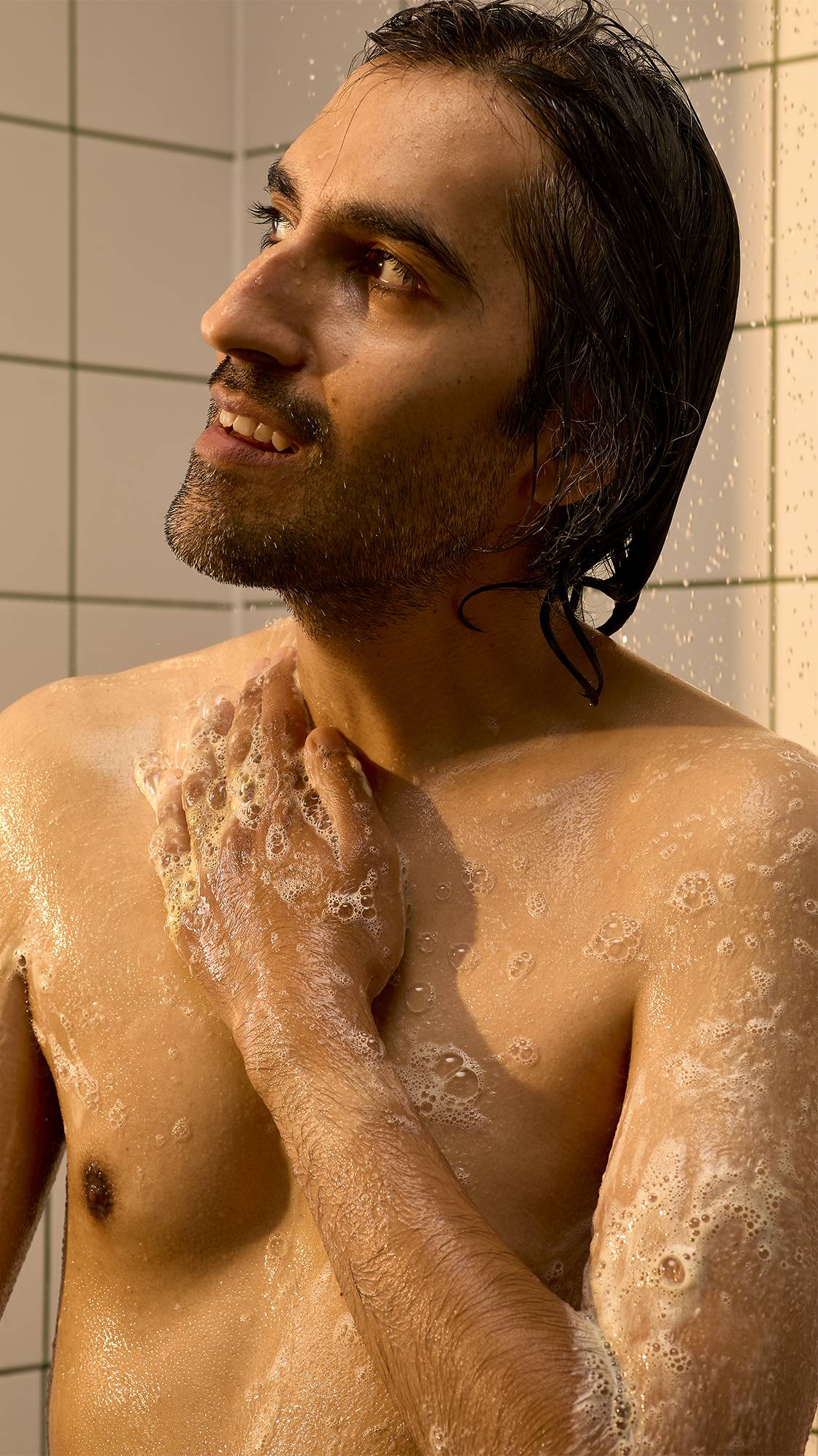 A model with a beard and wet hair smiles as they wash their chest and shoulders with foaming Lush "Mango Lassi" shower gel lather in a tiled shower setting.