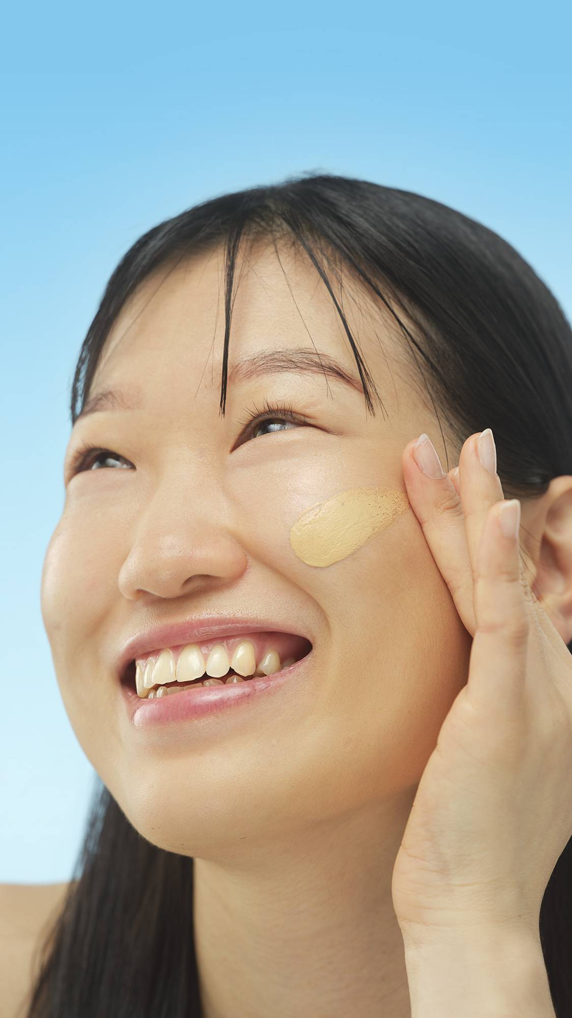 A person smiles brightly while applying a smooth, oval swatch of Medium Cool Skinwear foundation or skin tint to their cheek with a fingertip. They are looking upward against a light blue background.