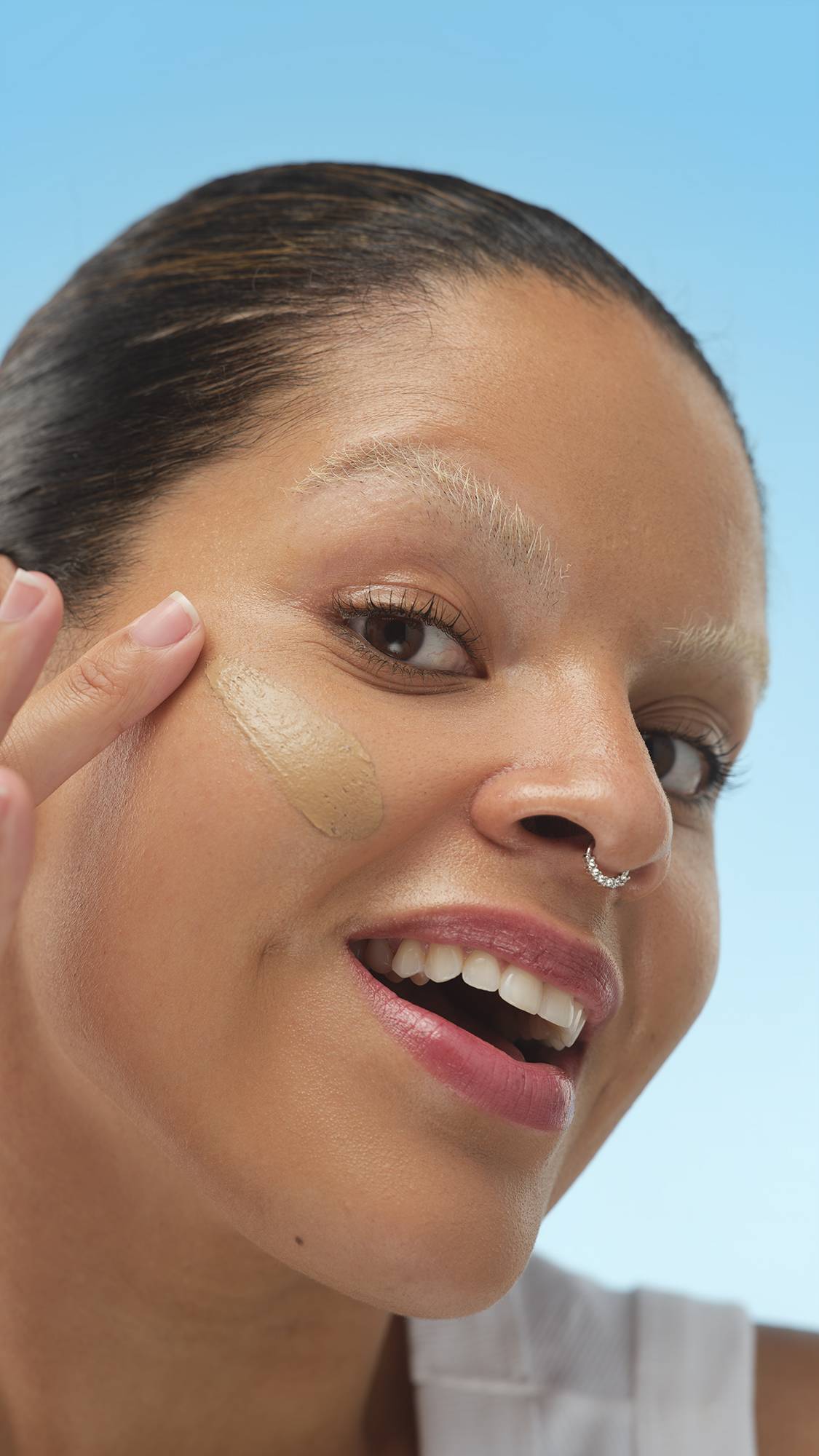 Smiling person with Medium Dark Cool Skinwear foundation, eyes closed, resting face in hands against a blue background. Features bleached eyebrows and a septum ring.