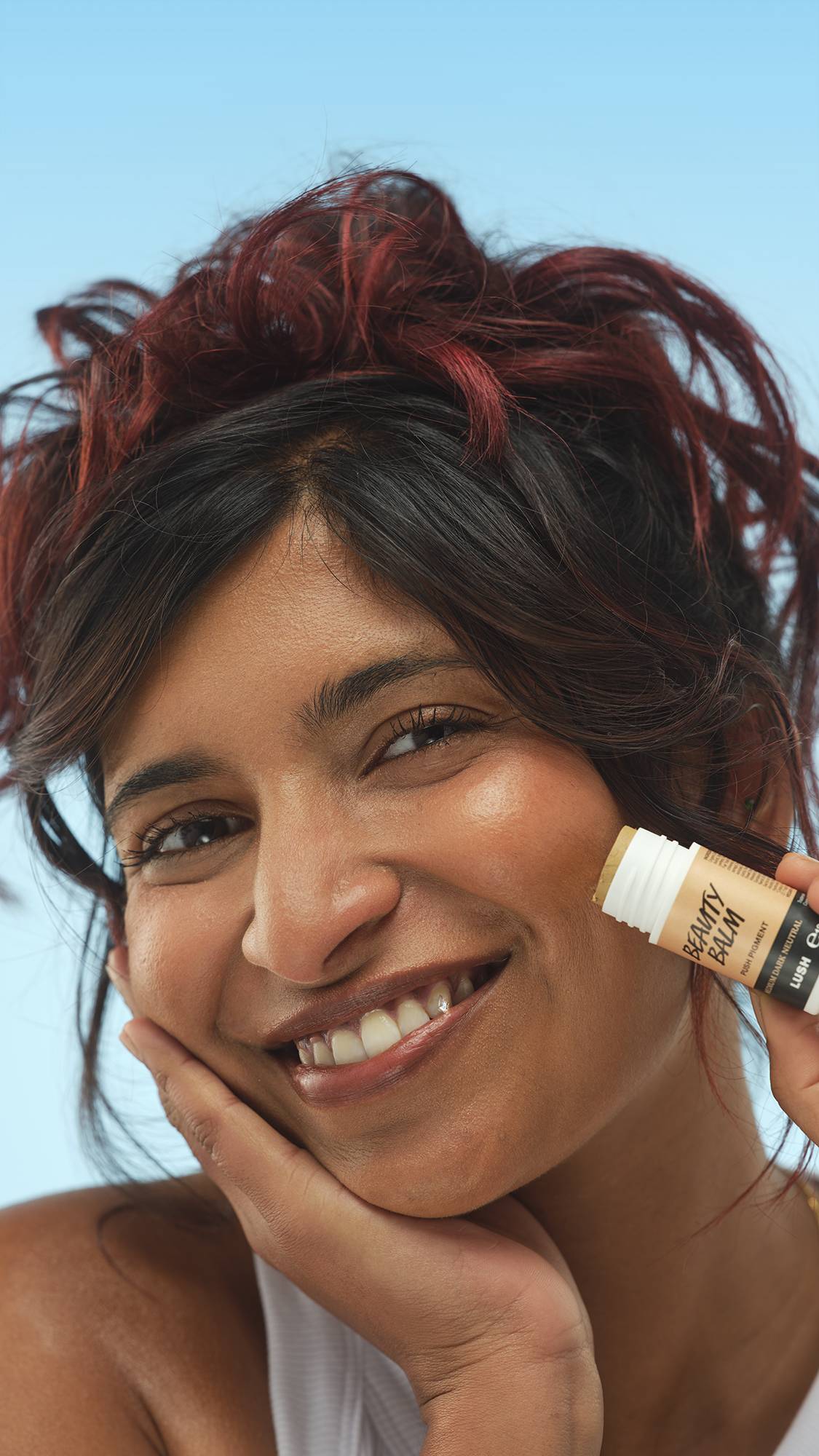 A close-up vertical portrait of a smiling person with, holding the stick of Medium Dark Neutral Beauty Balm to their cheek. Their hand is resting under their chin, with dark, red-highlighted hair, set against a bright blue background.