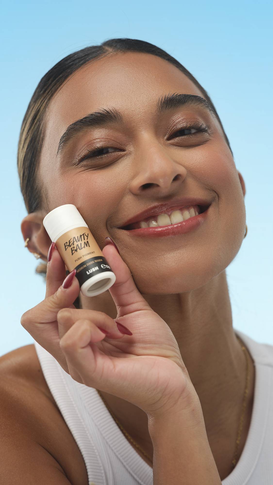 A close-up vertical portrait of a smiling person with medium-dark, warm-toned skin, holding the stick of Medium Dark Warm Beauty Balm to their cheek. They wear a white top and their hair is pulled back, set against a bright blue background