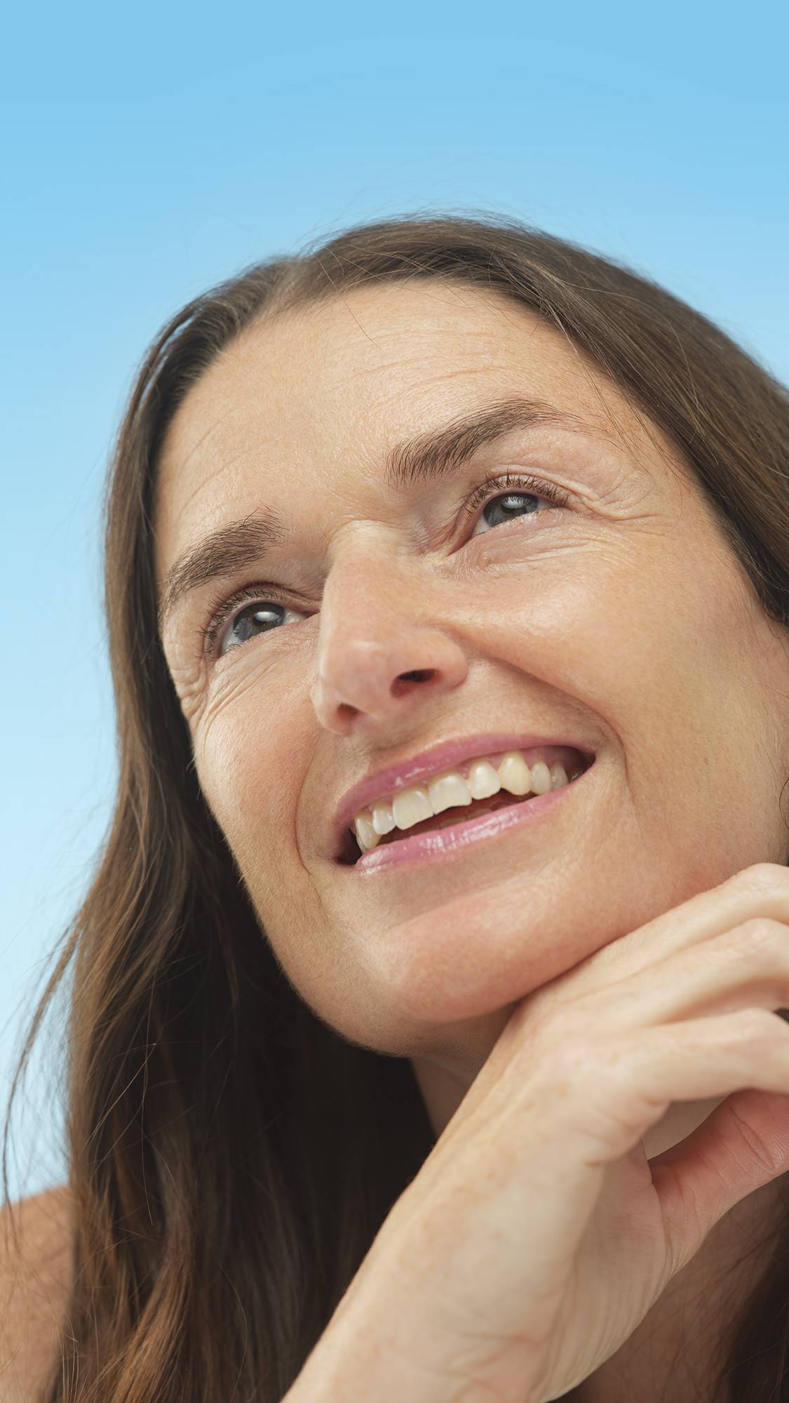 Close-up on a smiling person with Medium Neutral Skinwear foundation applied, looking upward with their chin resting on their hand against a light blue background.