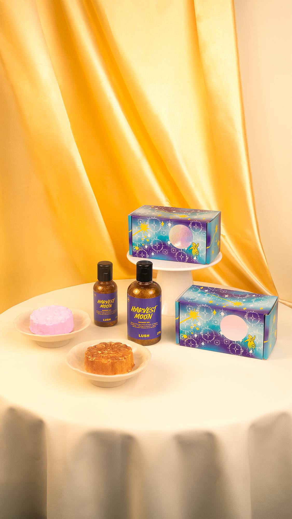 A display of Lush "Mid-Autumn Festival" products on a cream tablecloth against a draped gold backdrop. It features two starry purple gift boxes, two bottles of golden shower gel, a pink bubble bar, and a mooncake bath bomb.