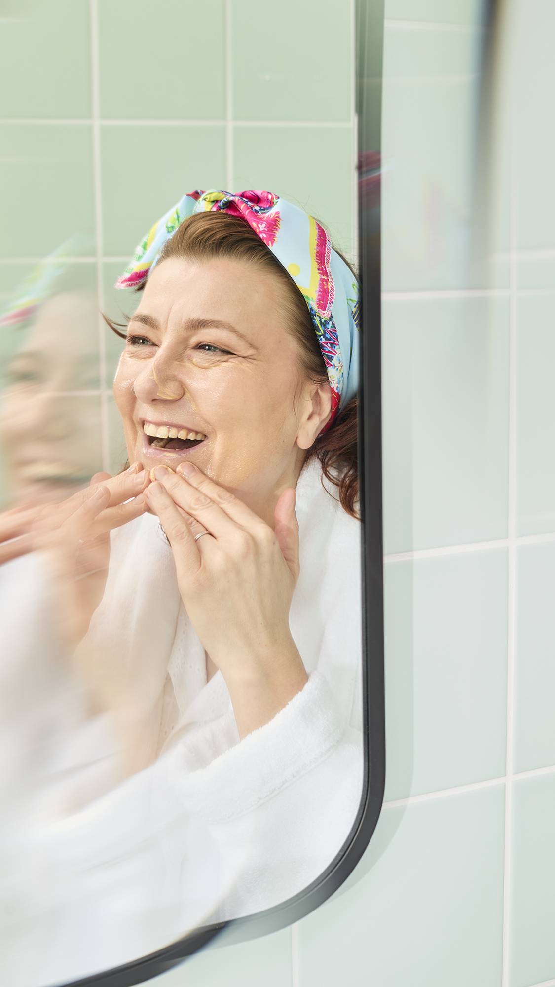 The image shows a model with wavy auburn hair in a bathroom mirror, smiling while applying a foundation with her hands. She has fashioned the colourful "My Family Houses" fabric knot wrap into a stylish headband.