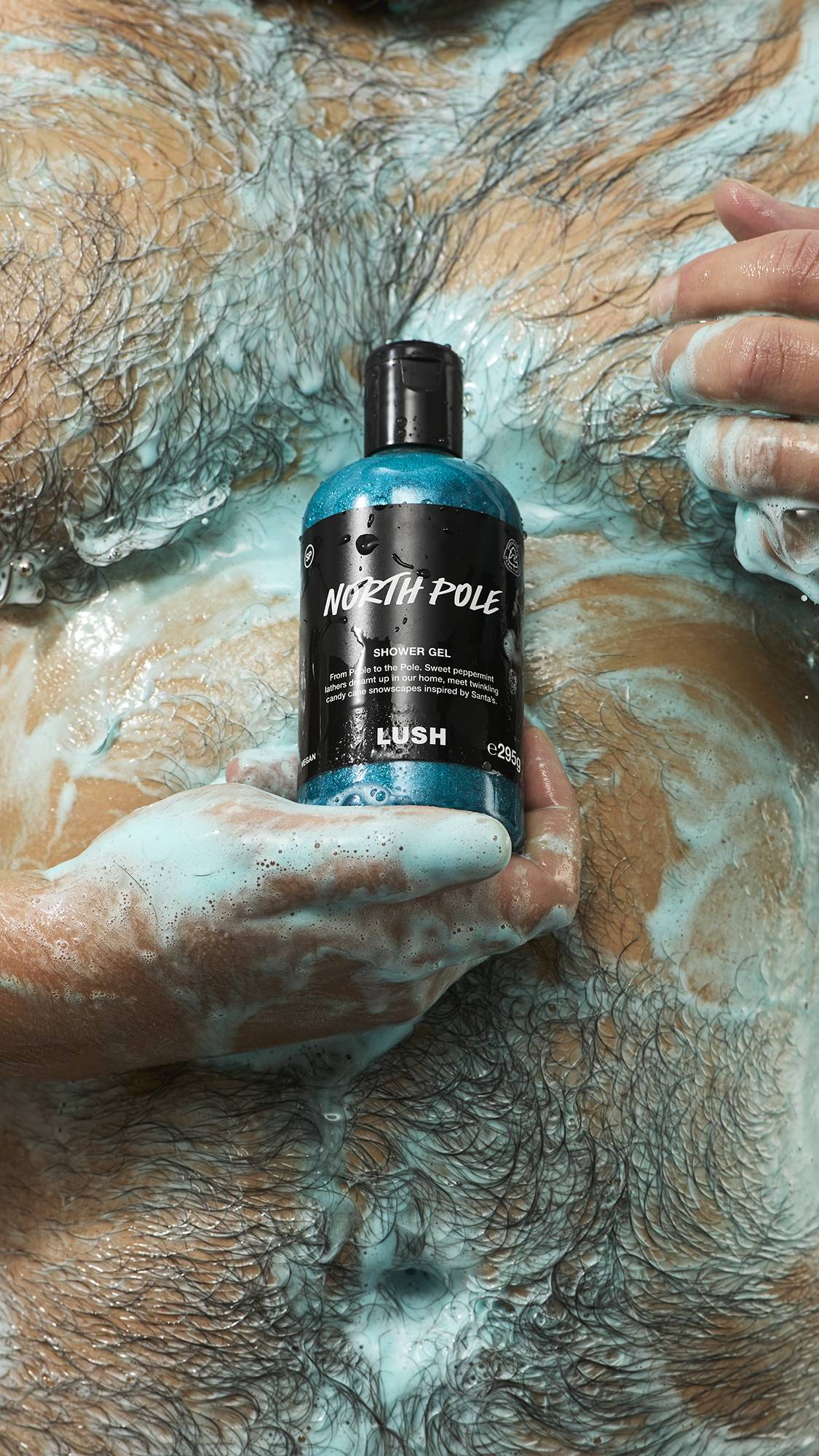 A soapy hand holds a bottle of Lush "North Pole" shimmering blue shower gel against a person's wet, hairy torso covered in light blue lather and froth.