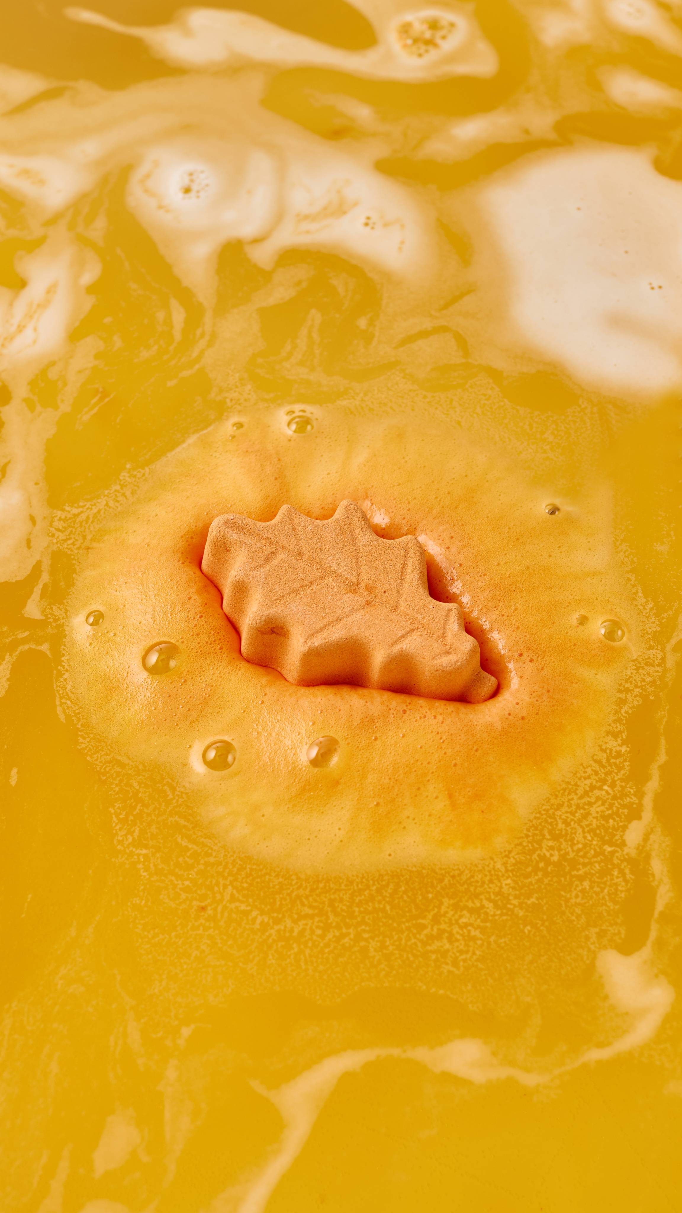 A top-down view of a small orange leaf-shaped bath bomb dissolving in the centre of soft, vibrant orange foam, resting atop swirling, warm golden-orange bathwater with light frothy patches.