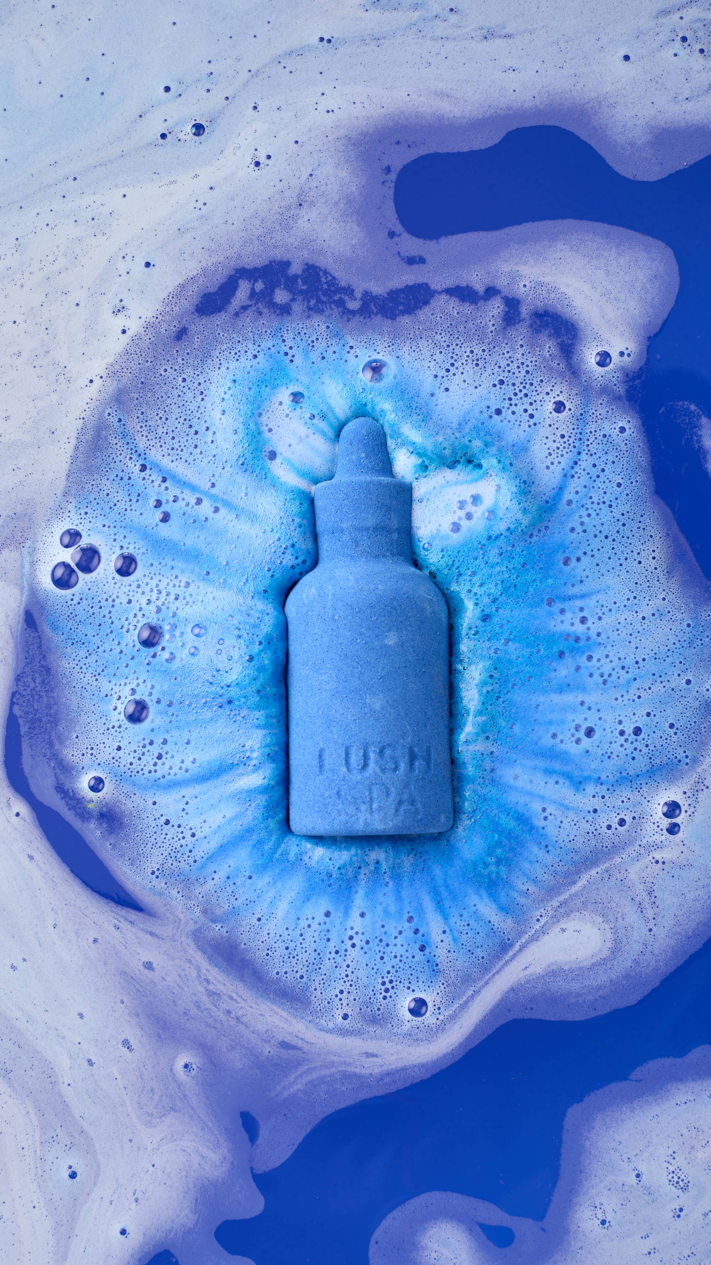 A vibrant blue 'Peace' bath bomb shaped like a dropper bottle dissolves in a bath of deep blue water. It is surrounded by a halo of bright blue and white fizz, with thick, frothy bubbles spreading across the surface of the water.