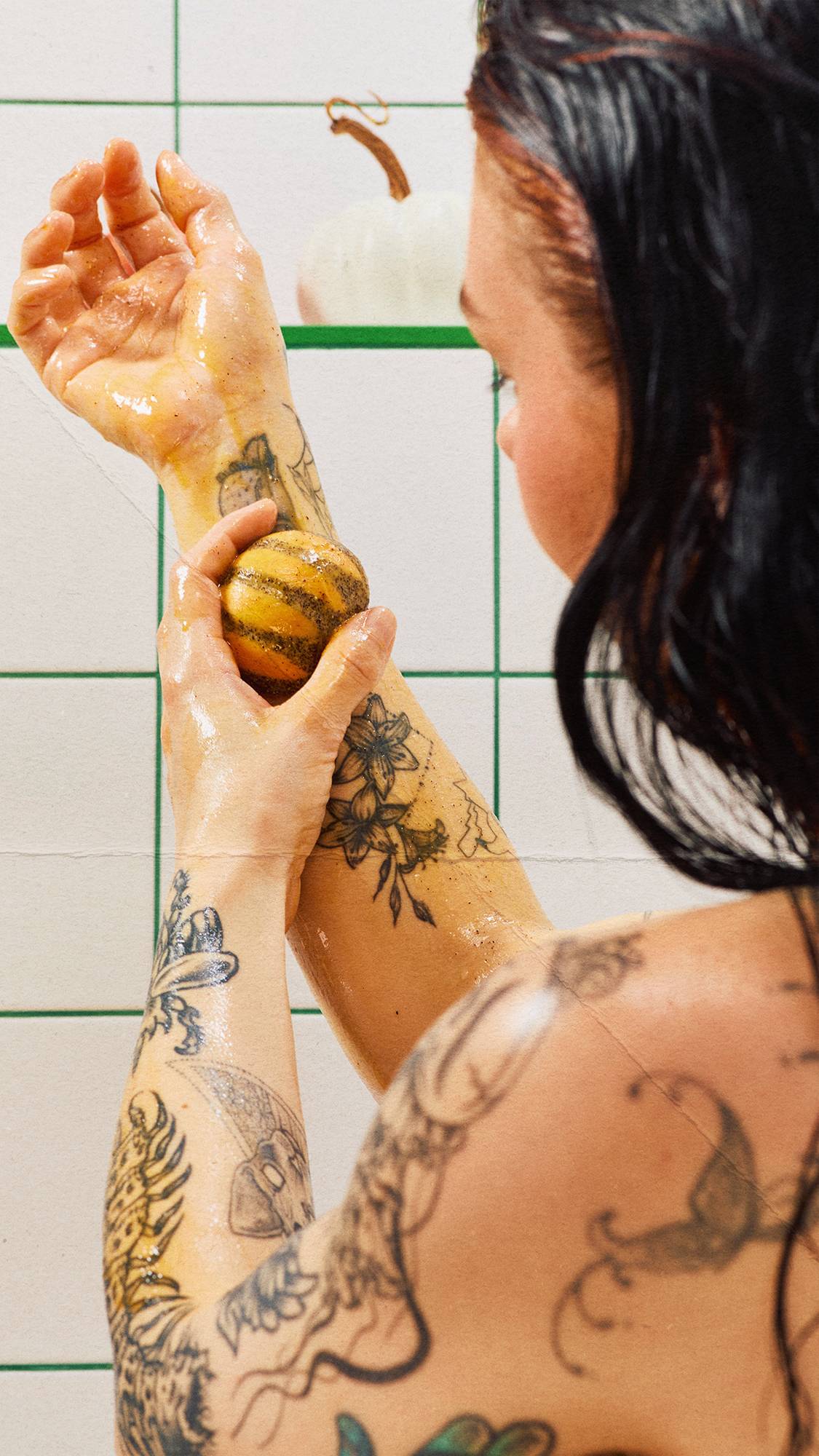 A tattooed person washes their forearm with an orange, pumpkin-shaped solid body scrub in the shower, set against white tiled walls with green grout and a white ornamental pumpkin on a ledge.
