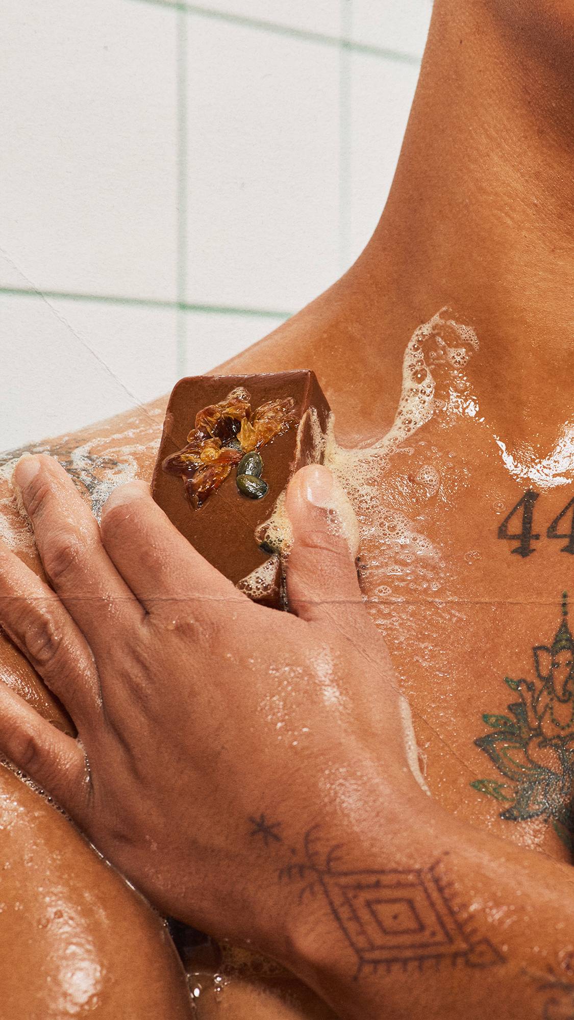 Close-up of a hand gliding a botanical-topped orange soap bar across a tattooed chest and shoulder, creating a white, bubbly lather in a green-grouted tiled shower.