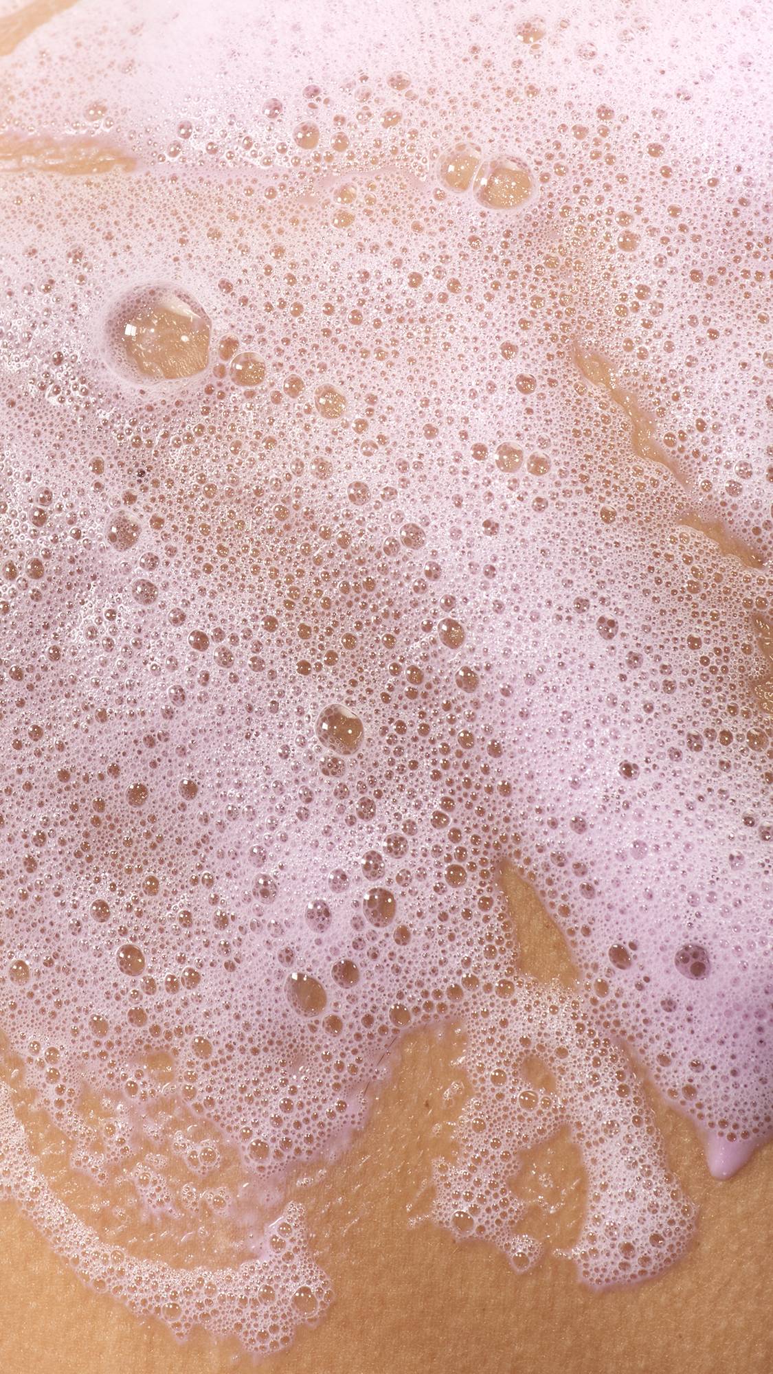 A close-up texture shot showing skin partially covered in a thick, bubbly layer of light pinkish-purple soap lather. Fine suds and larger translucent bubbles stretch across the surface.