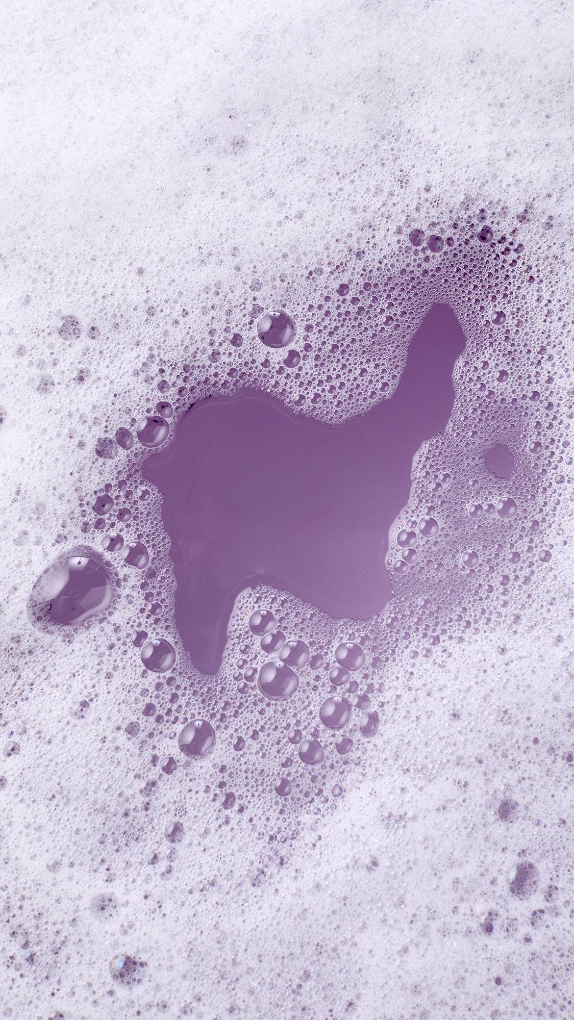 A thick layer of soft white bath foam parts to reveal deep lavender-purple bathwater beneath, surrounded by clusters of fine frothy bubbles.