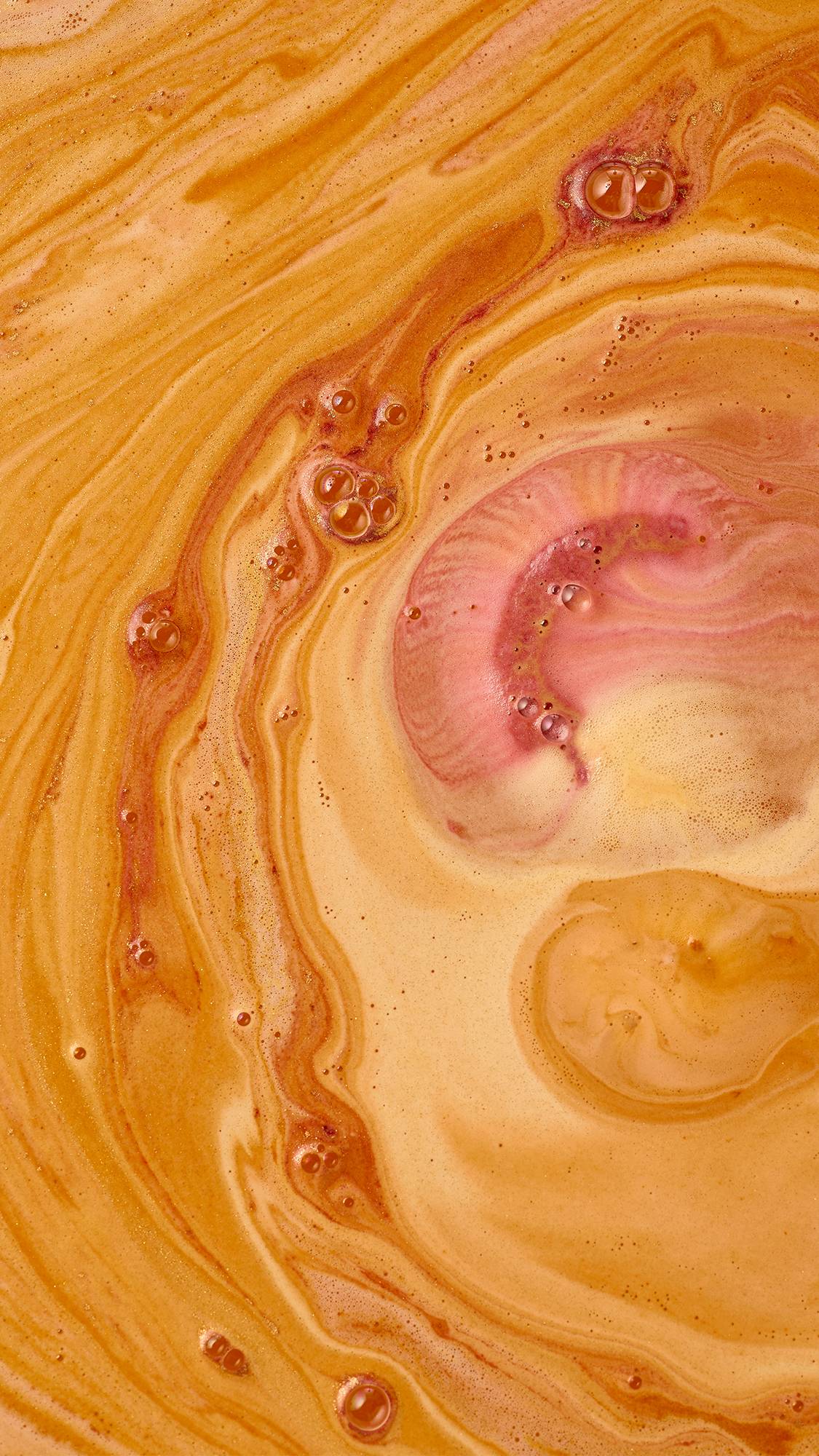 An overhead macro view of dissolving bath art, showing swirling patterns of rich golden-orange water mixed with creamy peach foam, fine lustrous shimmer, and soft pink streaks with scattered bubbles.