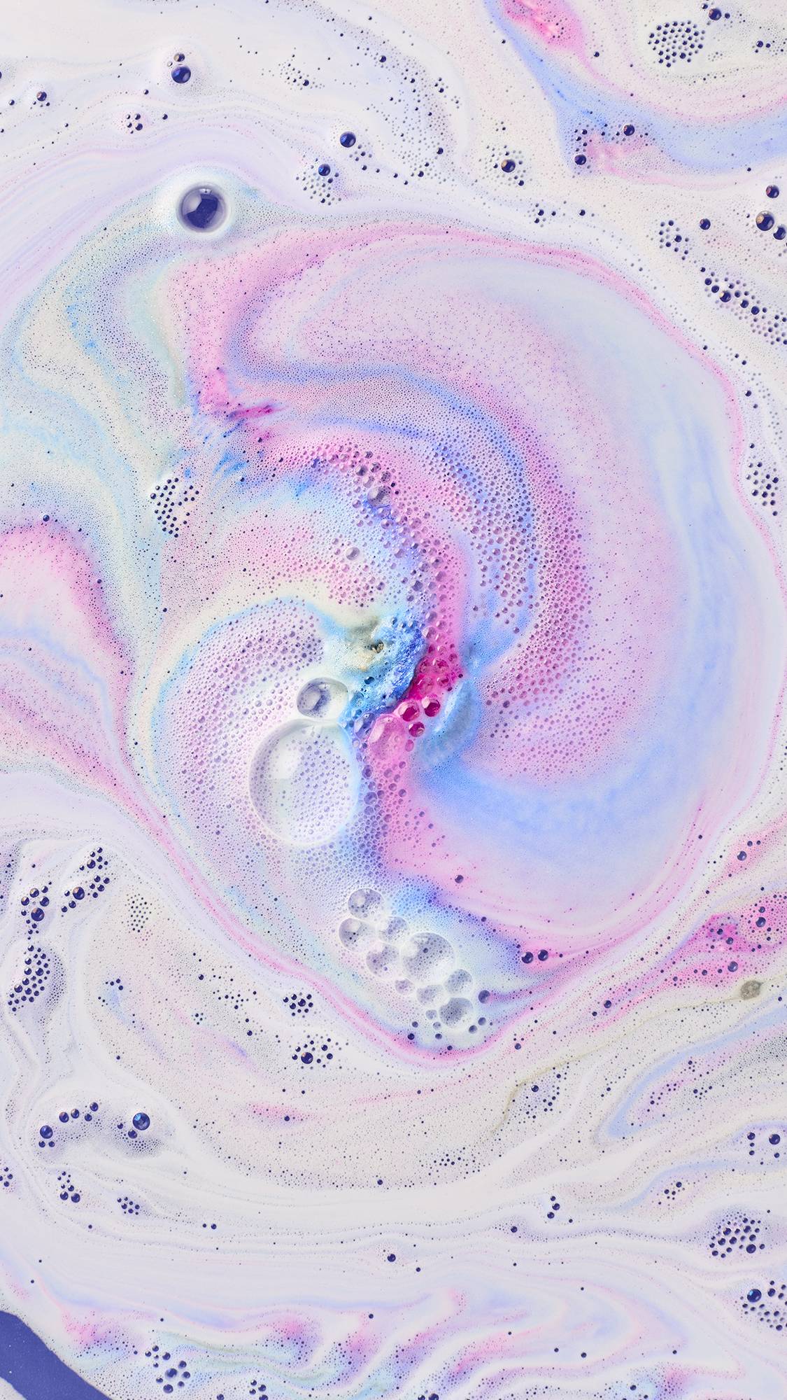 Soft swirls of lilac, periwinkle blue, and pastel pink bath foam spin across milky bathwater, with vibrant magenta accents and delicate bubbles floating at the centre.