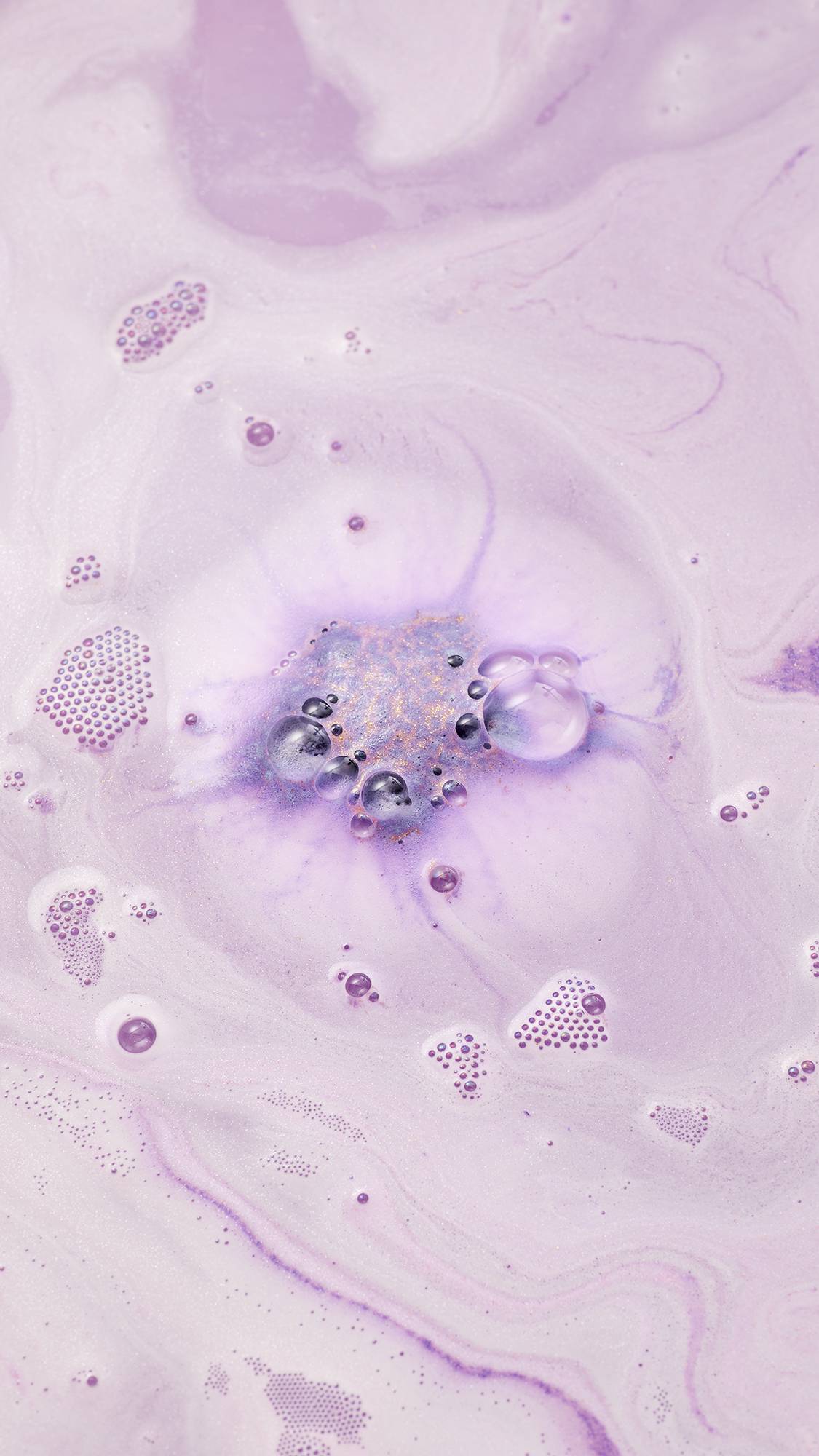 An overhead view of pale purple, foamy bath water. Clusters of white and purple bubbles float on the surface, while the center shows a concentrated swirl of deep purple and gold shimmer from the dissolved Siraj bath bomb.