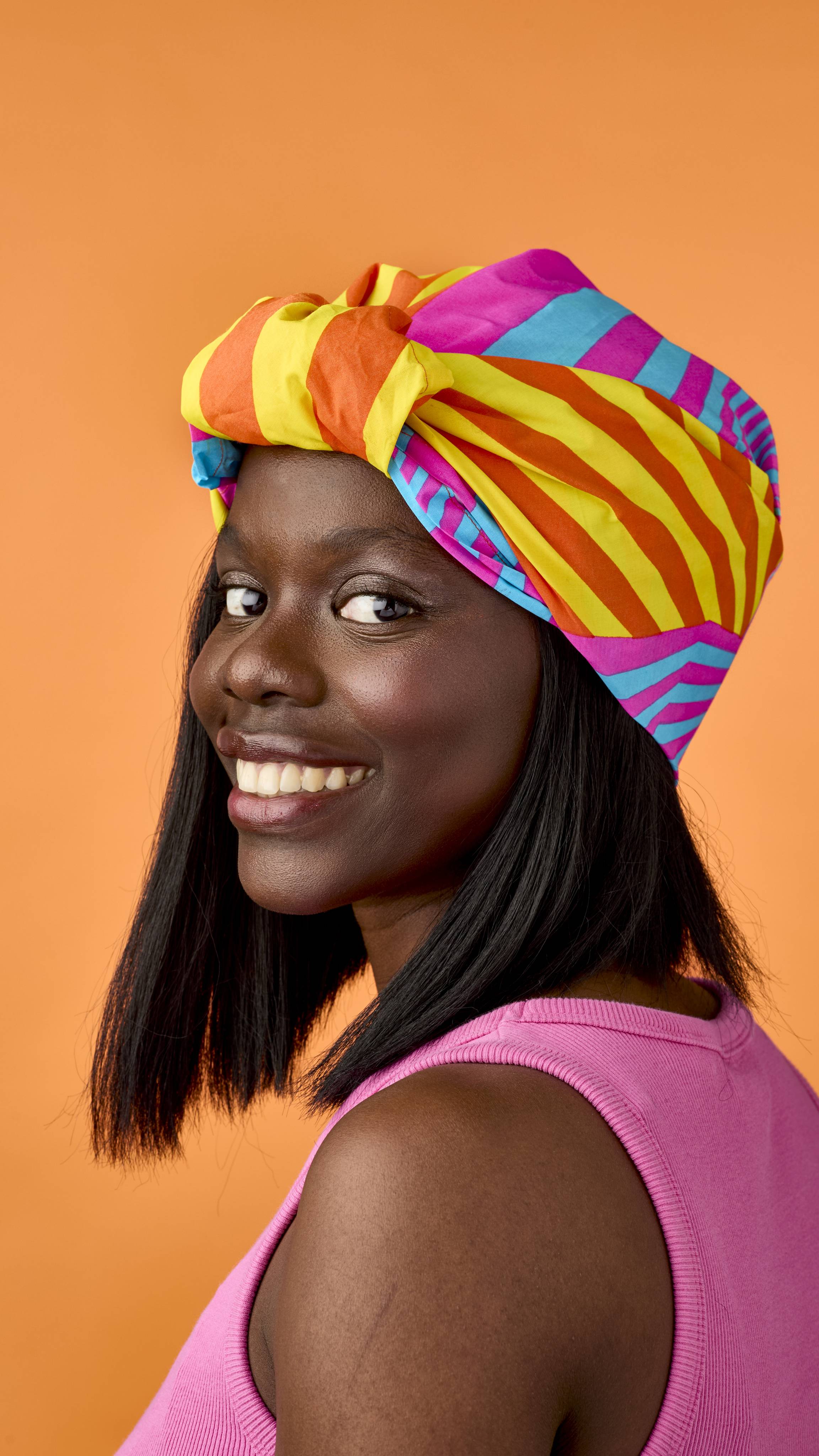 A smiling model in a pink top looks back over their shoulder, wearing a brightly coloured knot-wrap as a headwrap styled with yellow, orange, pink, and blue stripes against a warm orange backdrop.