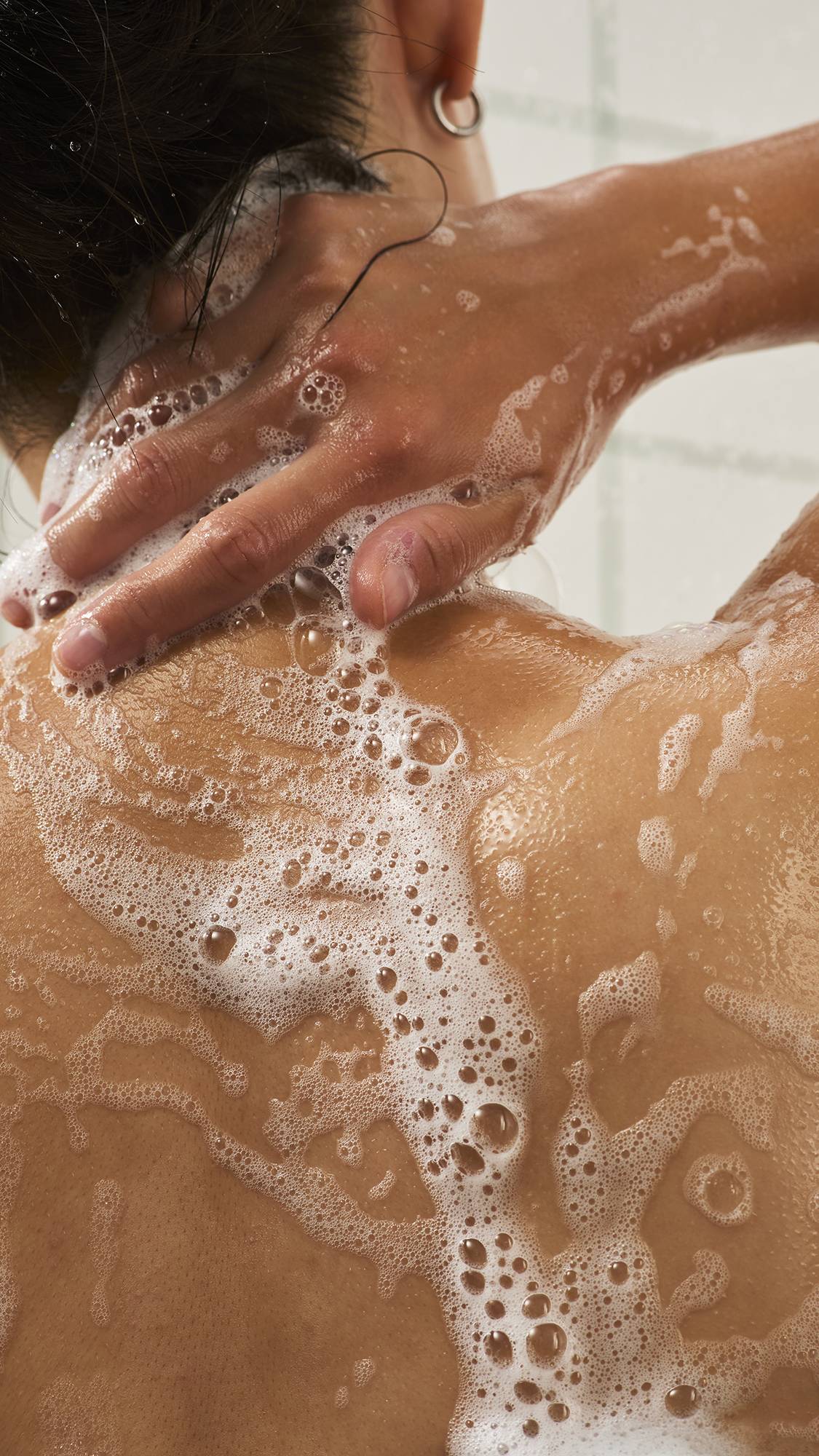 Close-up from behind of a person reaching back to wash the nape of their neck, with white soapy suds and frothy bubbles spread over their shoulders and back in the shower.
