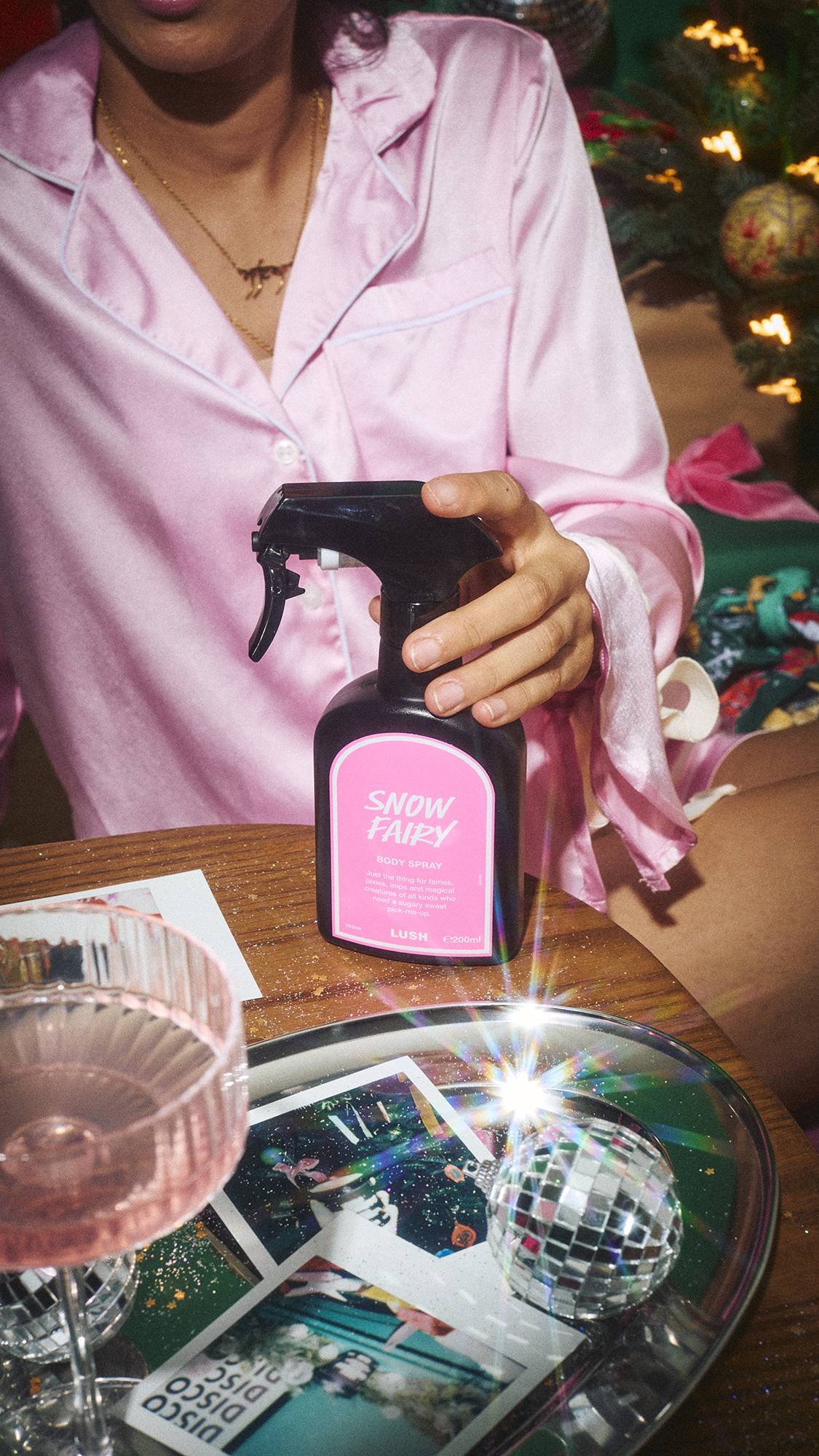 A close-up of a person in pink satin pyjamas holding a black spray bottle of Lush Snow Fairy body spray over a wooden table adorned with a silver tray, photos, a cocktail glass, and a mini disco ball.