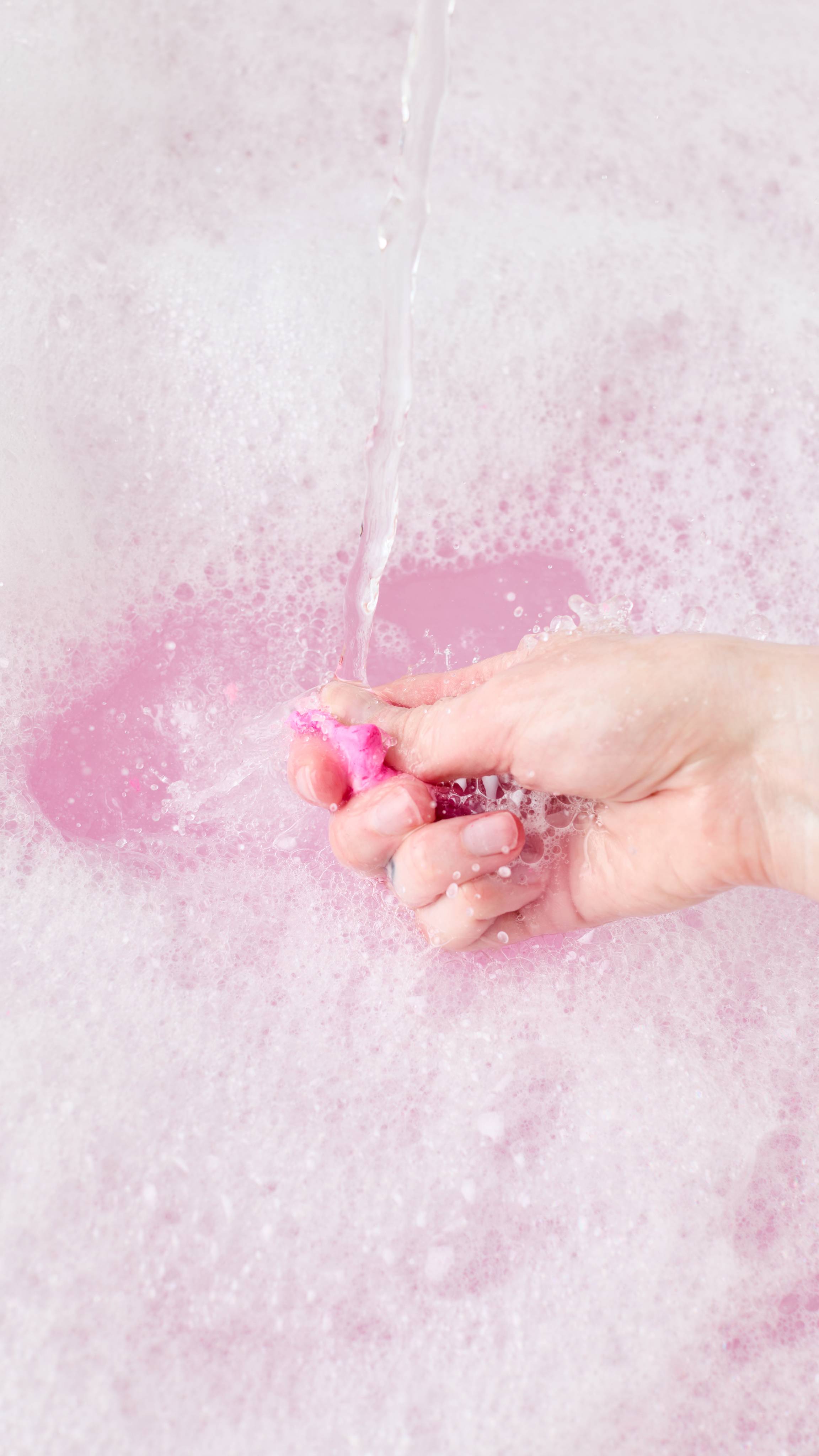 A hand crumbles some of the Lush "Snow Fairy" FUN product directly beneath a running stream of tap water, creating splash ripples over pastel pink bathwater covered in a sea of soft white bubbles.