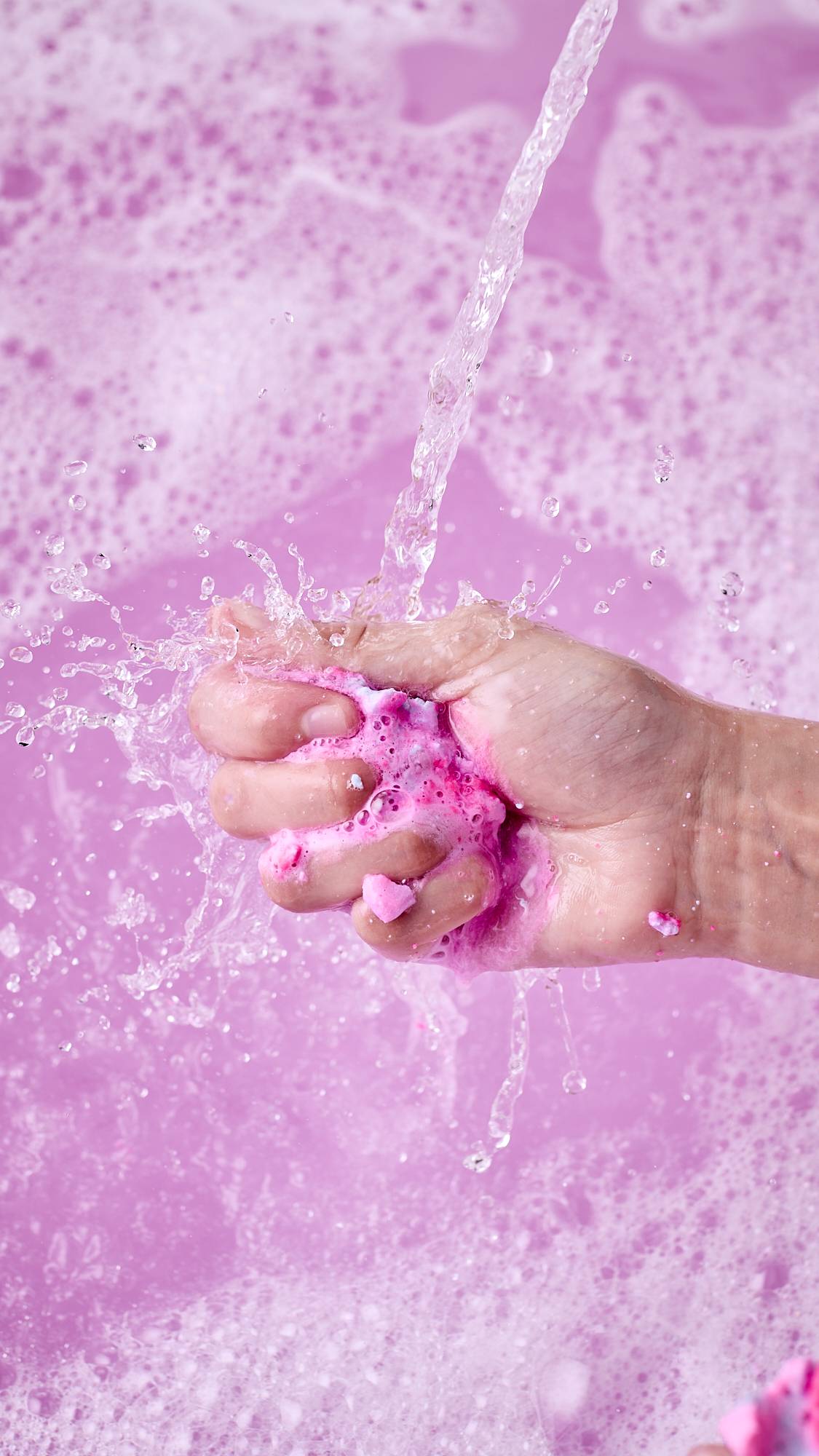 A close-up shot of a hand crushing a dissolving "Snowy Mooncake" bubble bar into a bright pink paste under a stream of running water. Water and pink suds splash over a bath filled with thick, frothy pink foam.