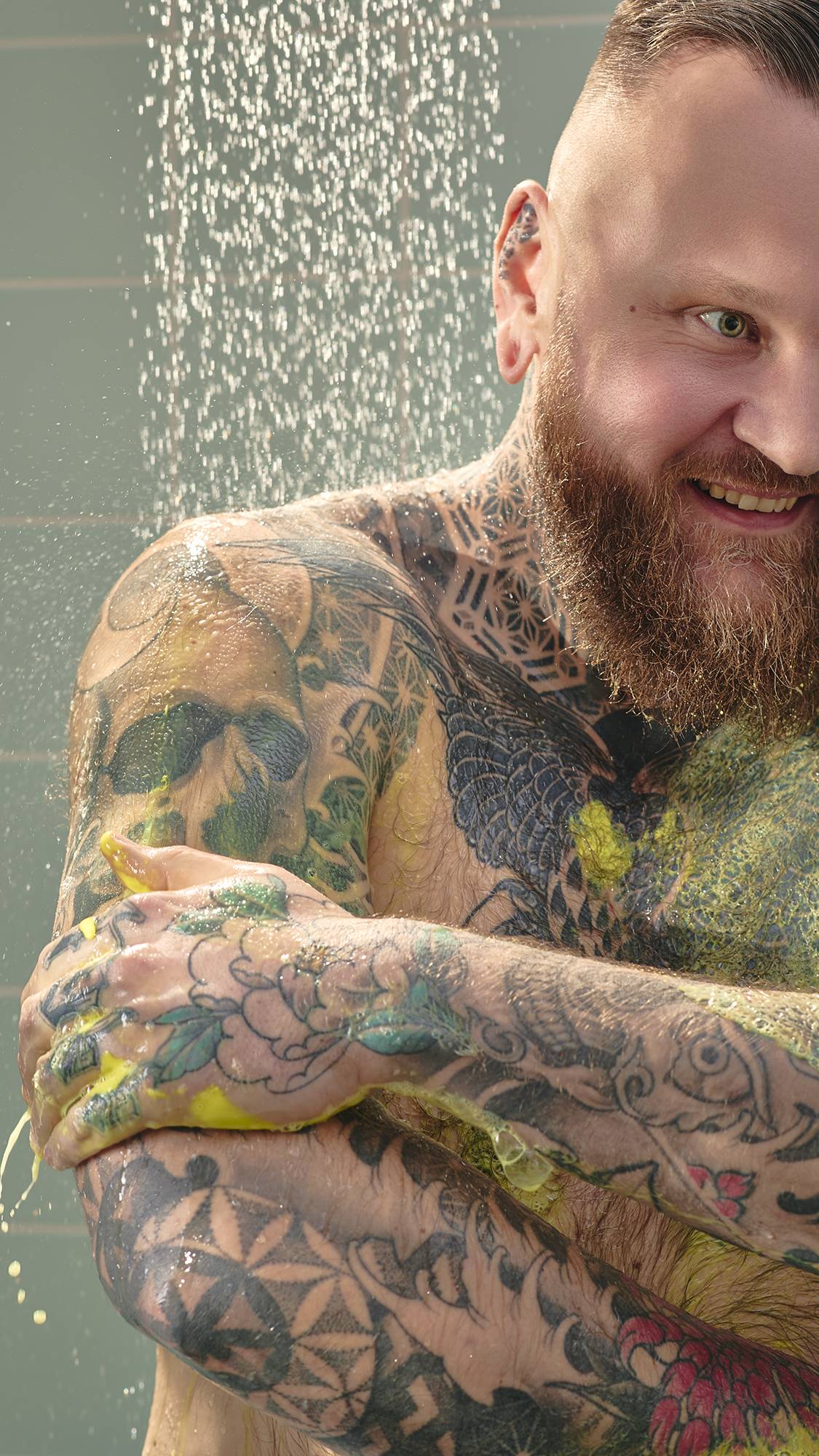 The image shows a model with a red-toned beard and extensive tattoos, smiling in the shower. They are lathering their arms with bright yellow "Squeeze The Day" shower gel, with water falling in the background against pale green tiles.