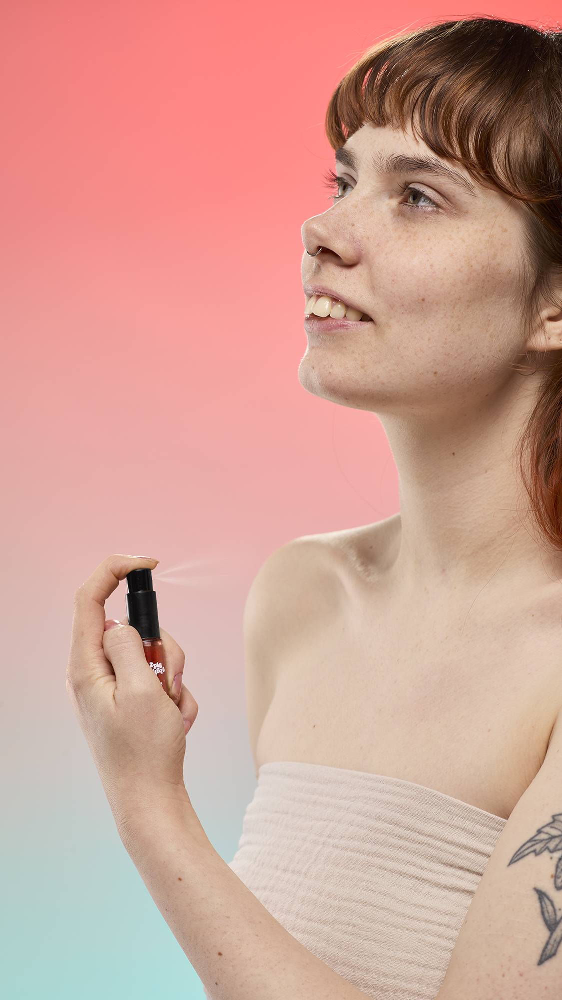 A person sprays a fine mist from a small reddish bottle of Little Squirts Sticky Dates Mini Glitter Mist toward their neck against a peach-to-blue background while wearing a light, textured wrap.