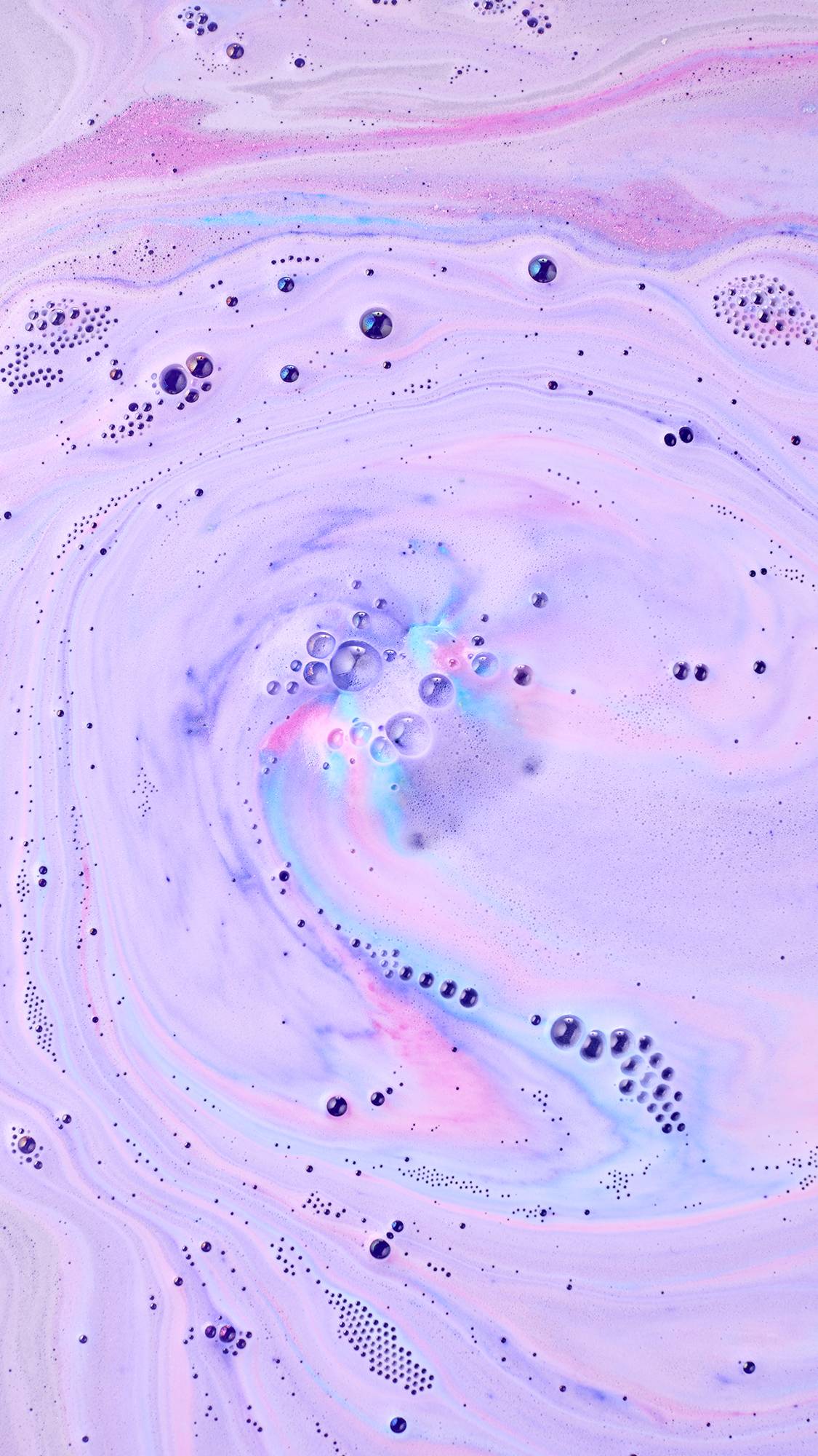 Swirling pastel lavender and white bathwater with dynamic streams of bright pink and cyan foam, dotted with scattered bubbles as a bath bomb fully dissolves.