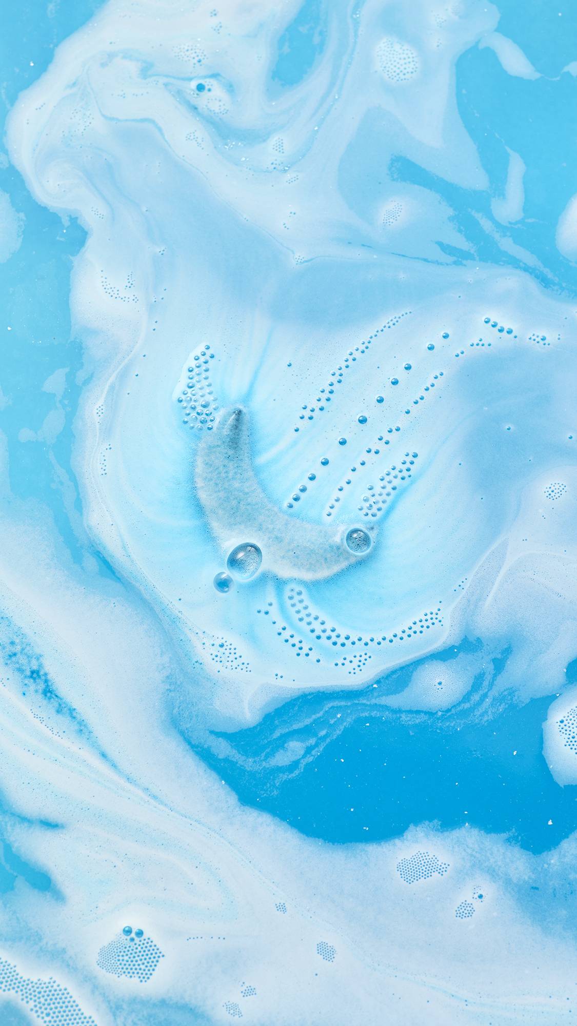An overhead view of the crescent moon-shaped Sukoon bath bomb dissolving in water. Swirls of vibrant blue and thick white foam surround the bath bomb, accompanied by delicate trails of small bubbles on the surface.