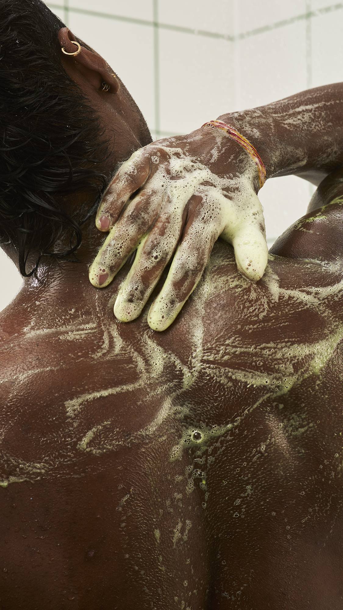A person seen from behind in the shower, reaching back to massage soft greenish-white soap lather across their neck and shoulder against a white tiled wall.