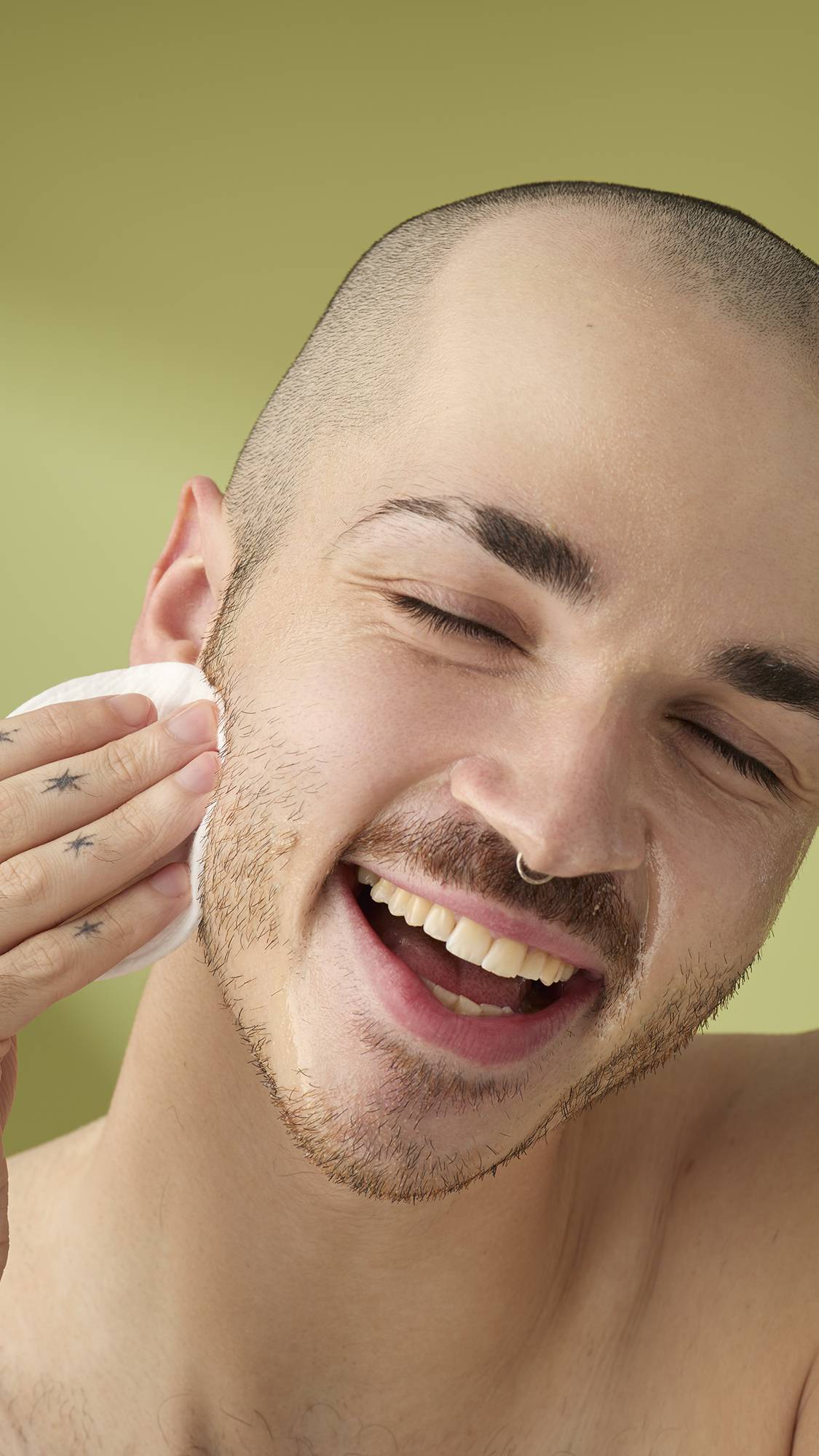 A smiling person with a shaved head and facial hair applies Ultra Plant to their cheek using a cotton pad. They have star tattoos on their fingers and closed eyes against a green background.