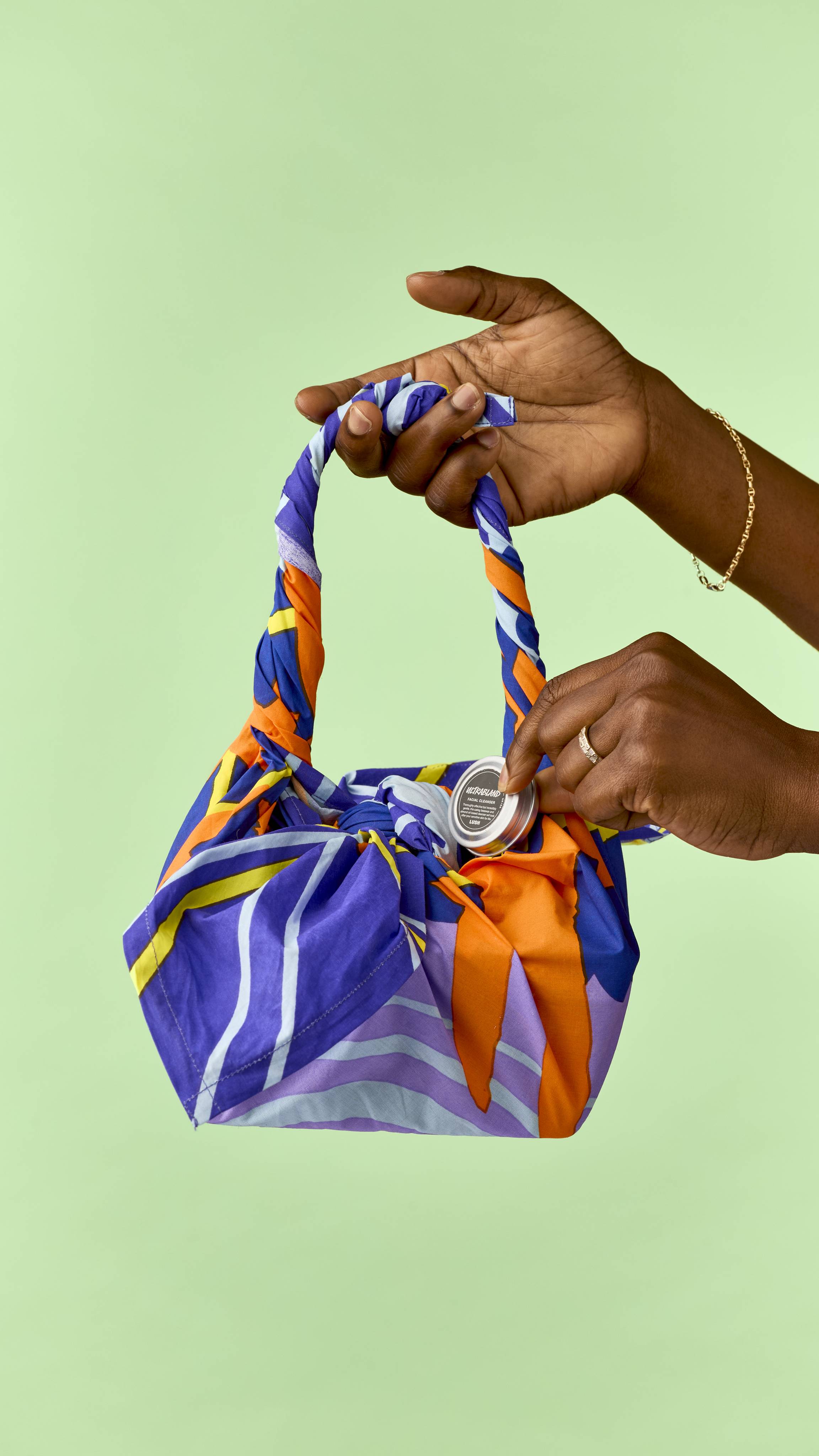 Hands place a small silver tin inside a knot-wrapped fabric bag styled with a tied handle in purple, orange, blue, and yellow patterns, held against a solid light green background.