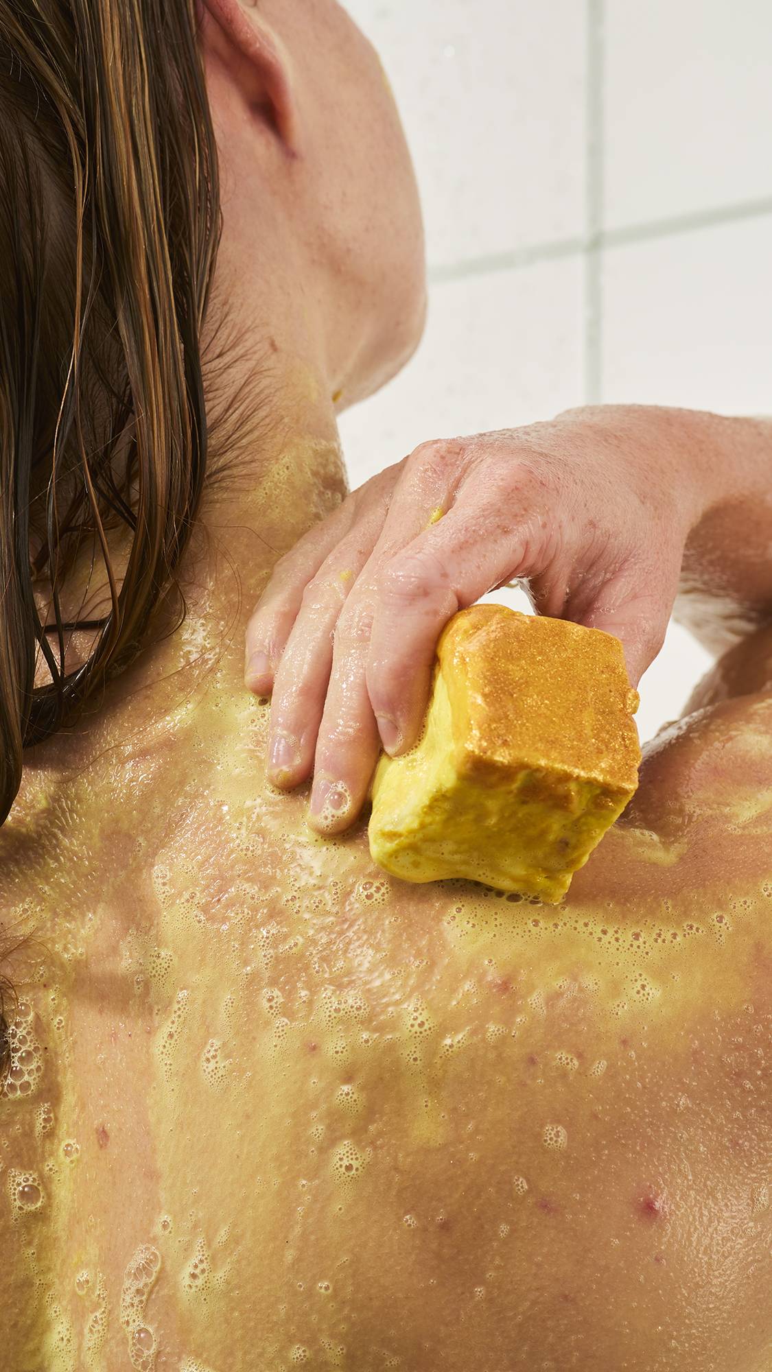 A person rubs a yellow cube soap bar directly onto their shoulder in the shower, spreading creamy yellow suds over wet skin against a white tiled background.