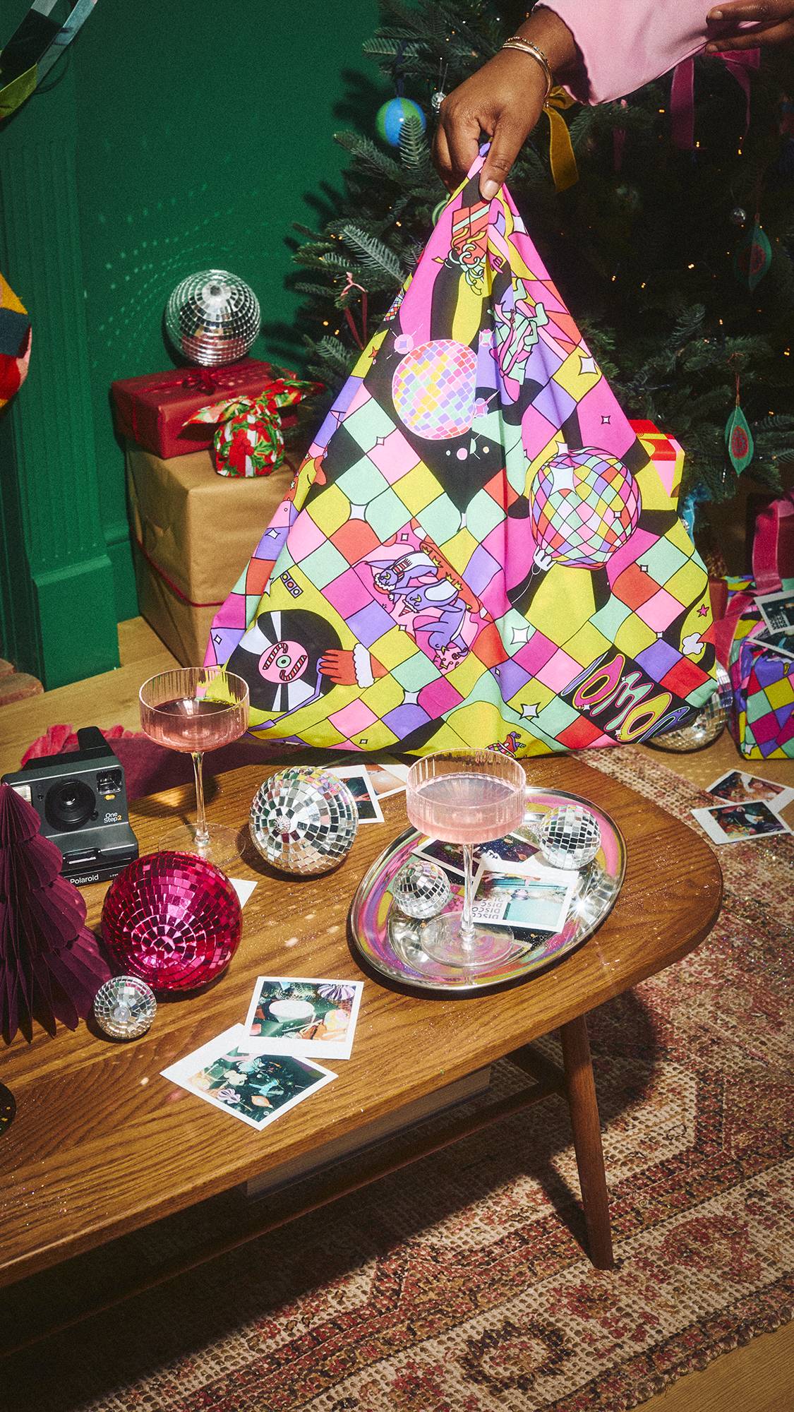 A hand holds up a gift wrapped in a colourful, festive disco-patterned Lush Knot Wrap like a bag, set in a party room with a coffee table holding drinks, disco balls, and instant photos, near a decorated Christmas tree.