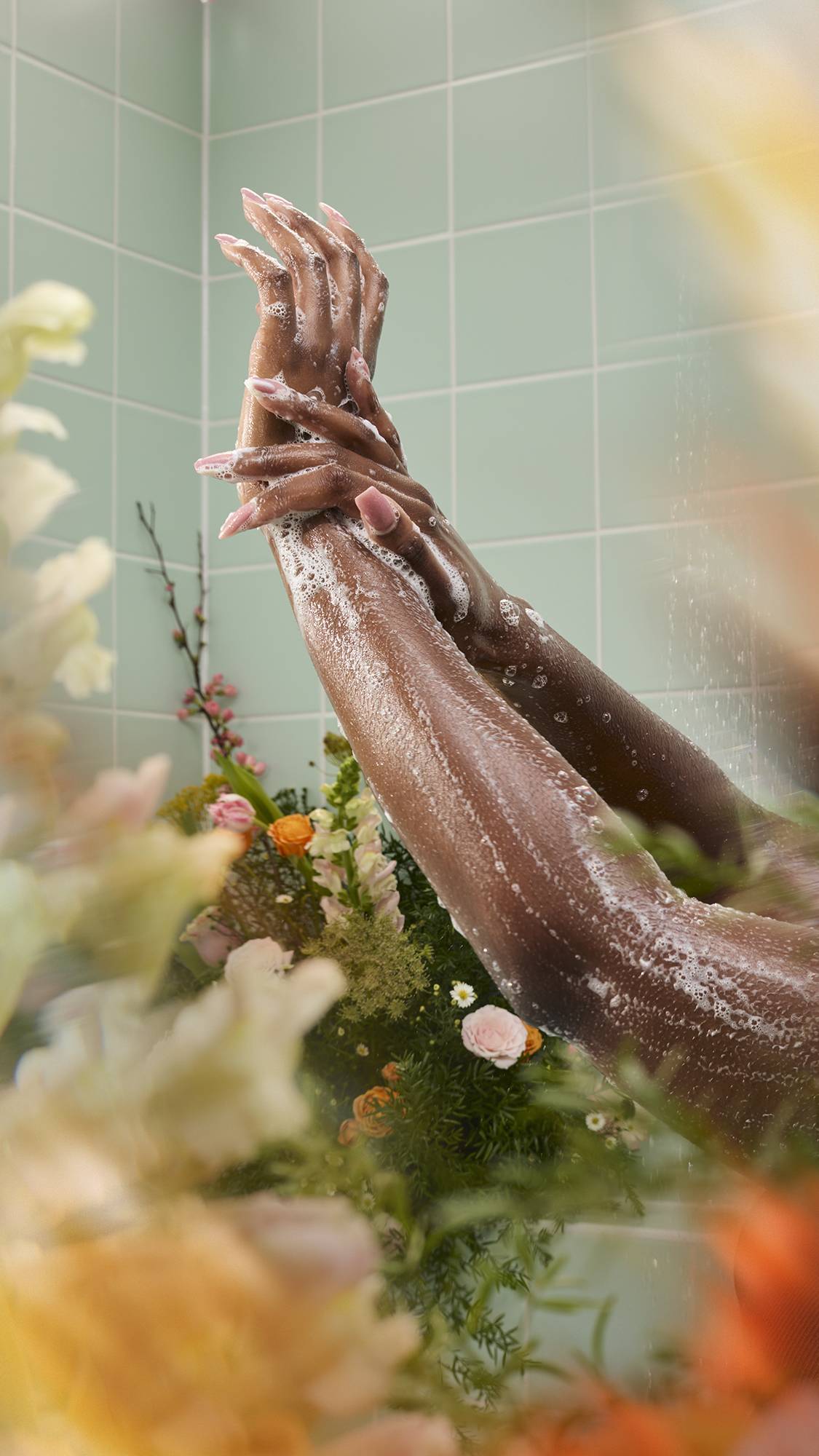 Two wet arms reach upward, covered in a light, soapy lather from the "Yummy Custard" body wash. The shower scene is framed by a lush, blurred arrangement of flowers in the foreground, set against a backdrop of mint green tiles.