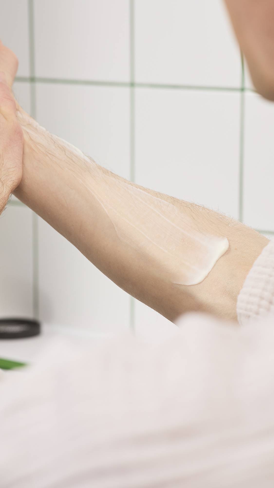 A hand smoothes rich, cream-coloured Lush "Yuzu Cheesecake" body butter along a forearm, creating a thick, moisturising layer against a white tiled bathroom background.