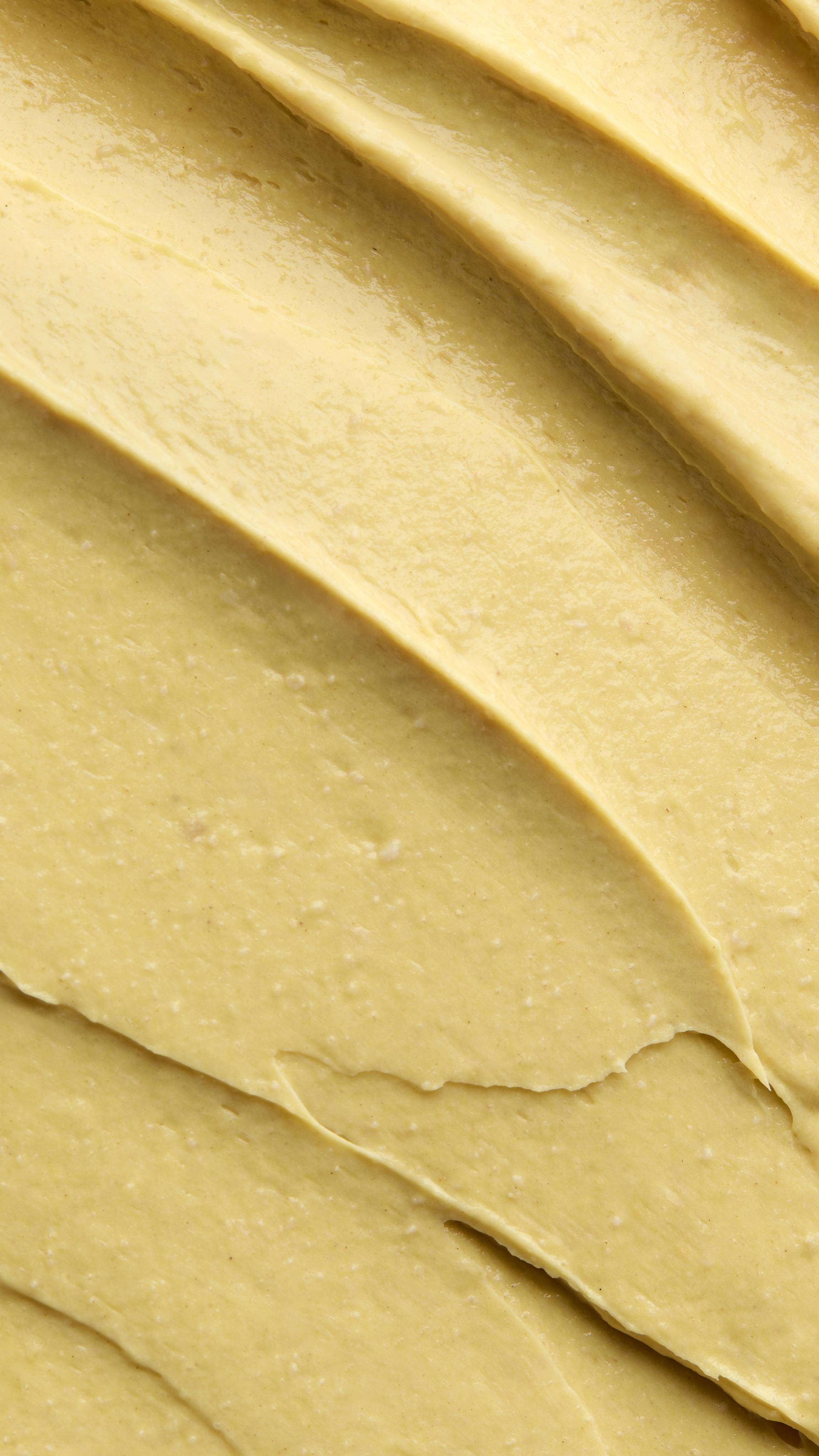 A close-up, textured shot of a thick, custard-yellow "Ahaa" scalp treatment spread in diagonal ridges. The product has a creamy yet grainy consistency with visible fine white specks throughout.