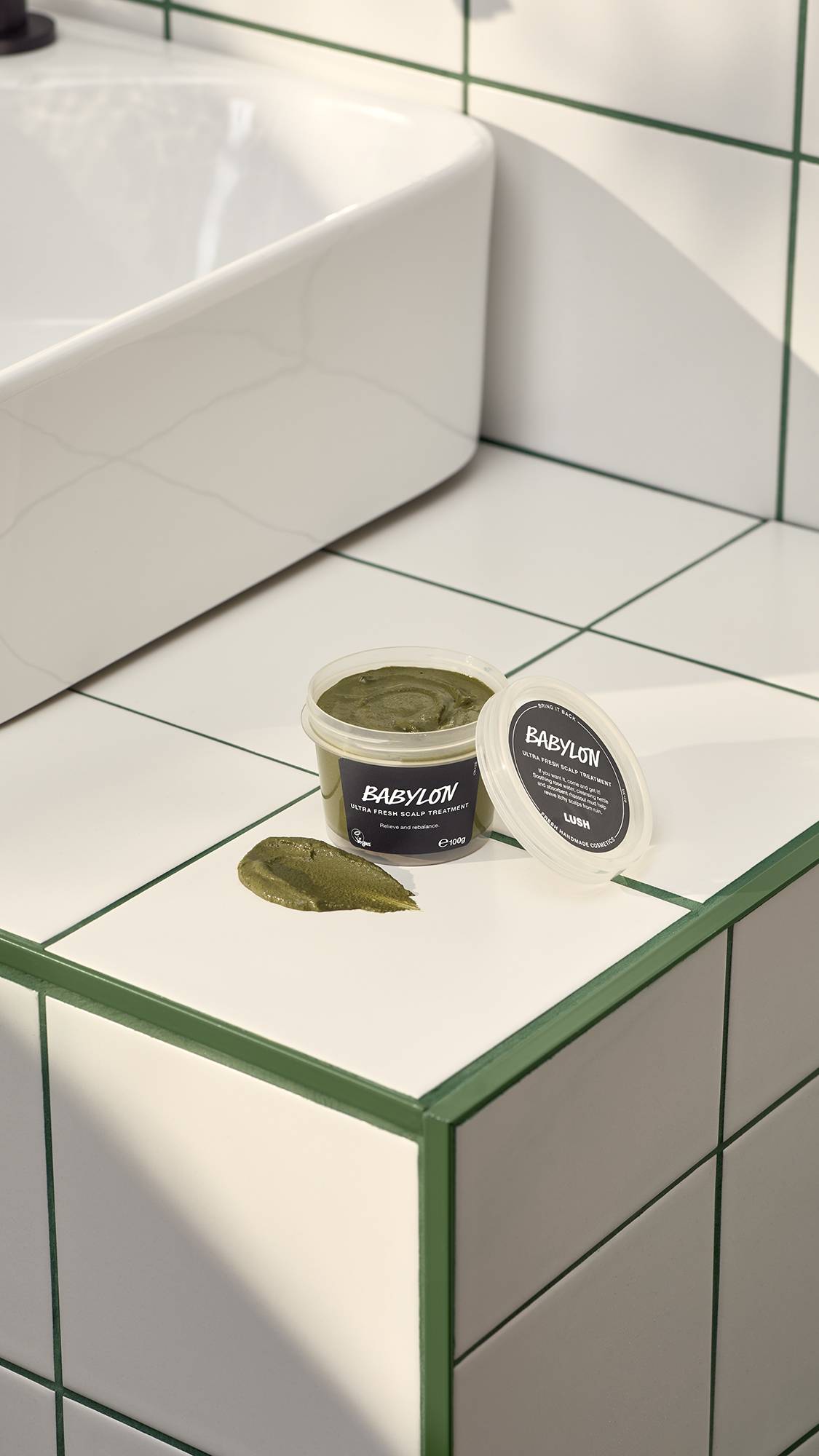 An open pot of Lush "Babylon" fresh scalp treatment sits on a white tiled surface with green grout next to its lid. A thick smear of the dark olive-green, grainy paste rests on the tile. A white sink is visible in the background.