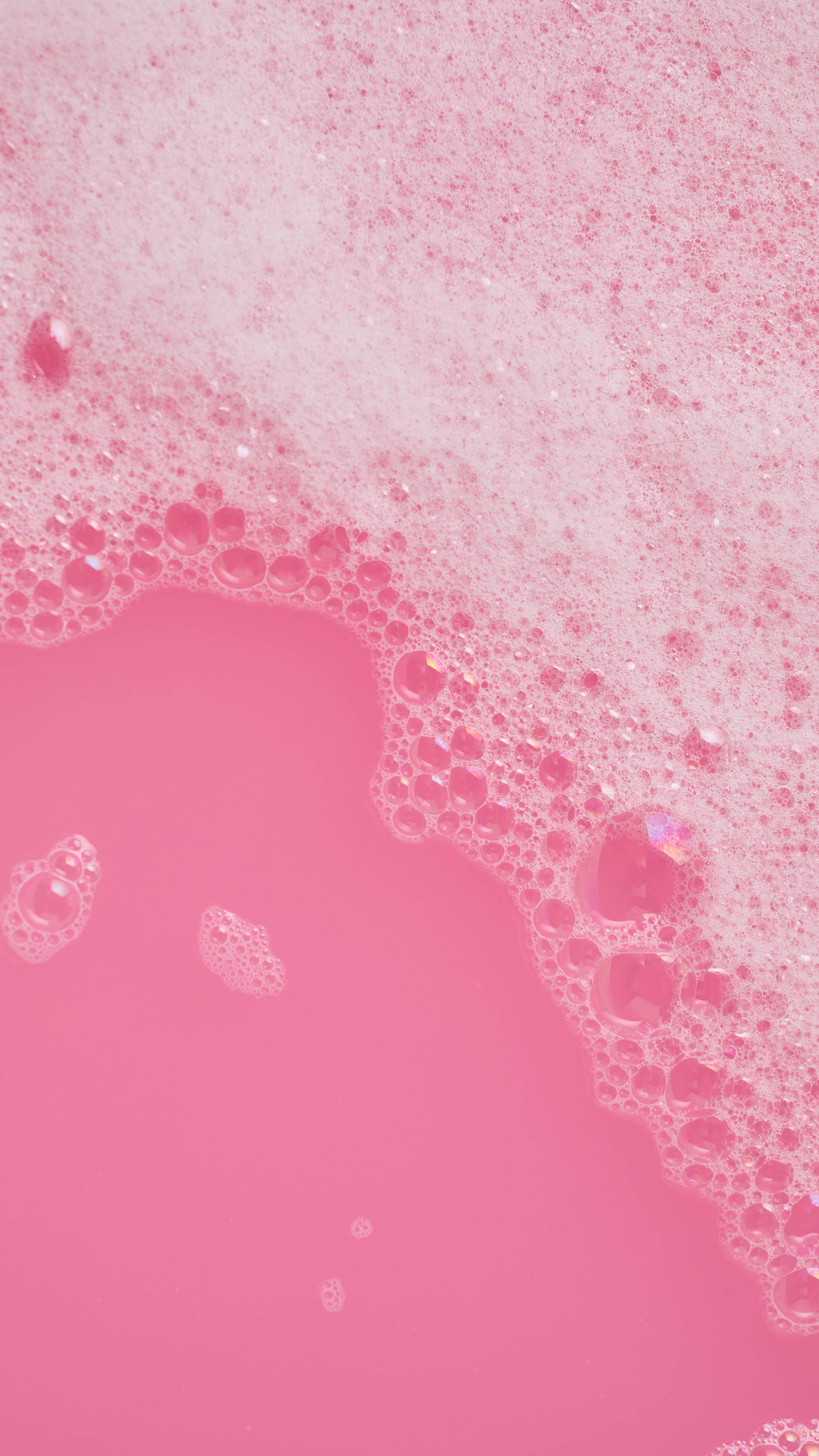 A thick, fluffy blanket of white and soft pink bath foam drifts across vibrant bubblegum-pink water, creating fine shimmering bubbles along the edge.