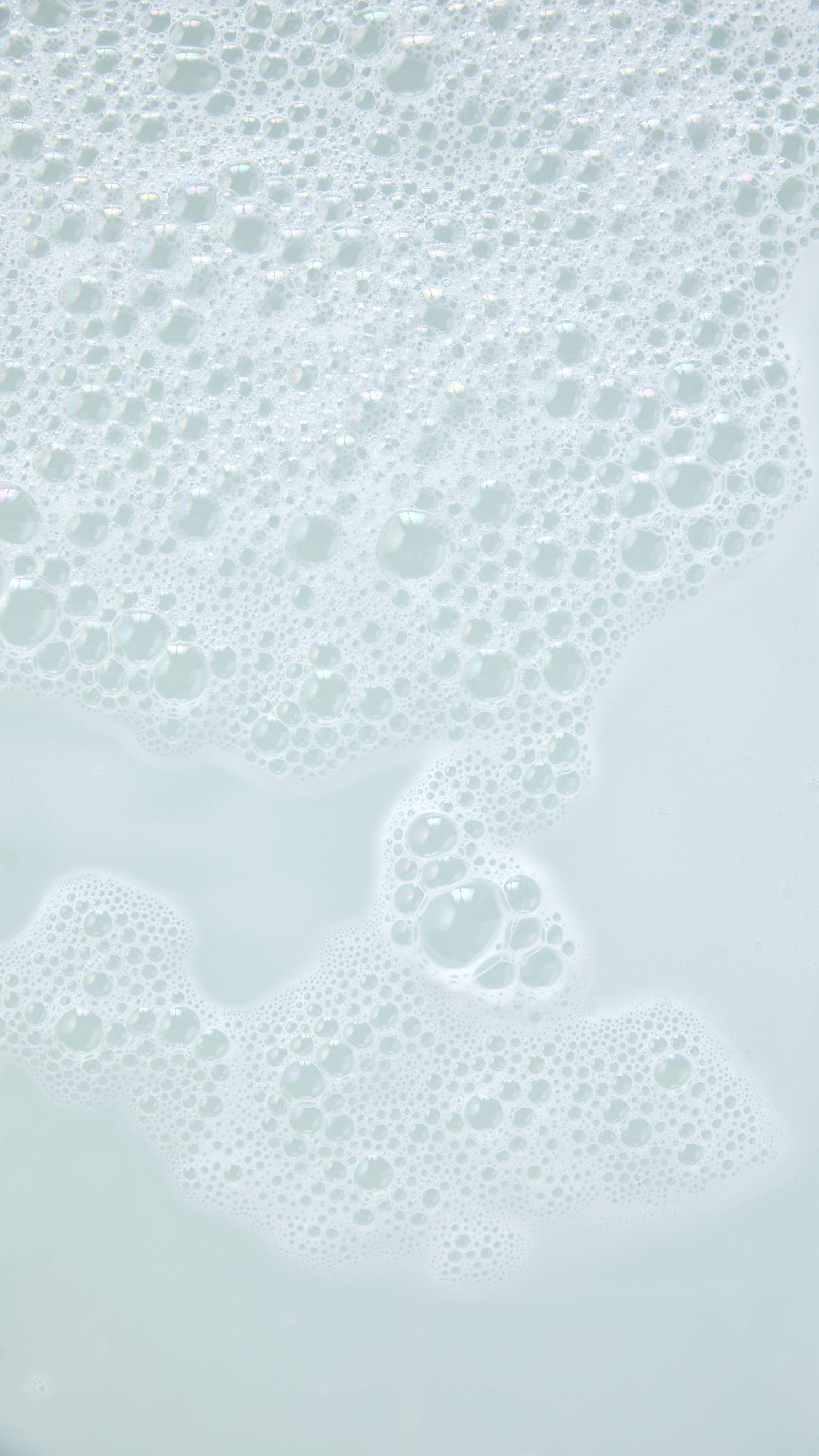 A thick, fluffy layer of soft white bath foam drifts across pale, milky white bathwater, creating clusters of fine, iridescent bubbles.