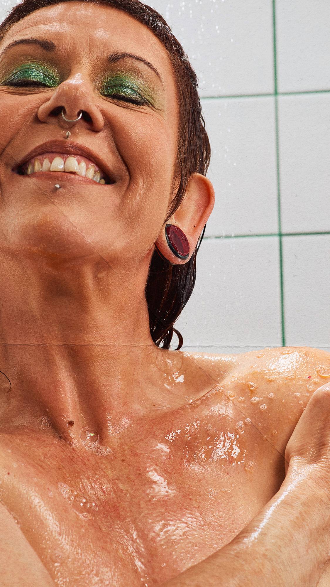 Close-up of a smiling person with green eyeshadow, facial piercings, and ear gauges, enjoying a shower as water and bubbly shower gel lather wash over their shoulders against a white-tiled wall with green grout.