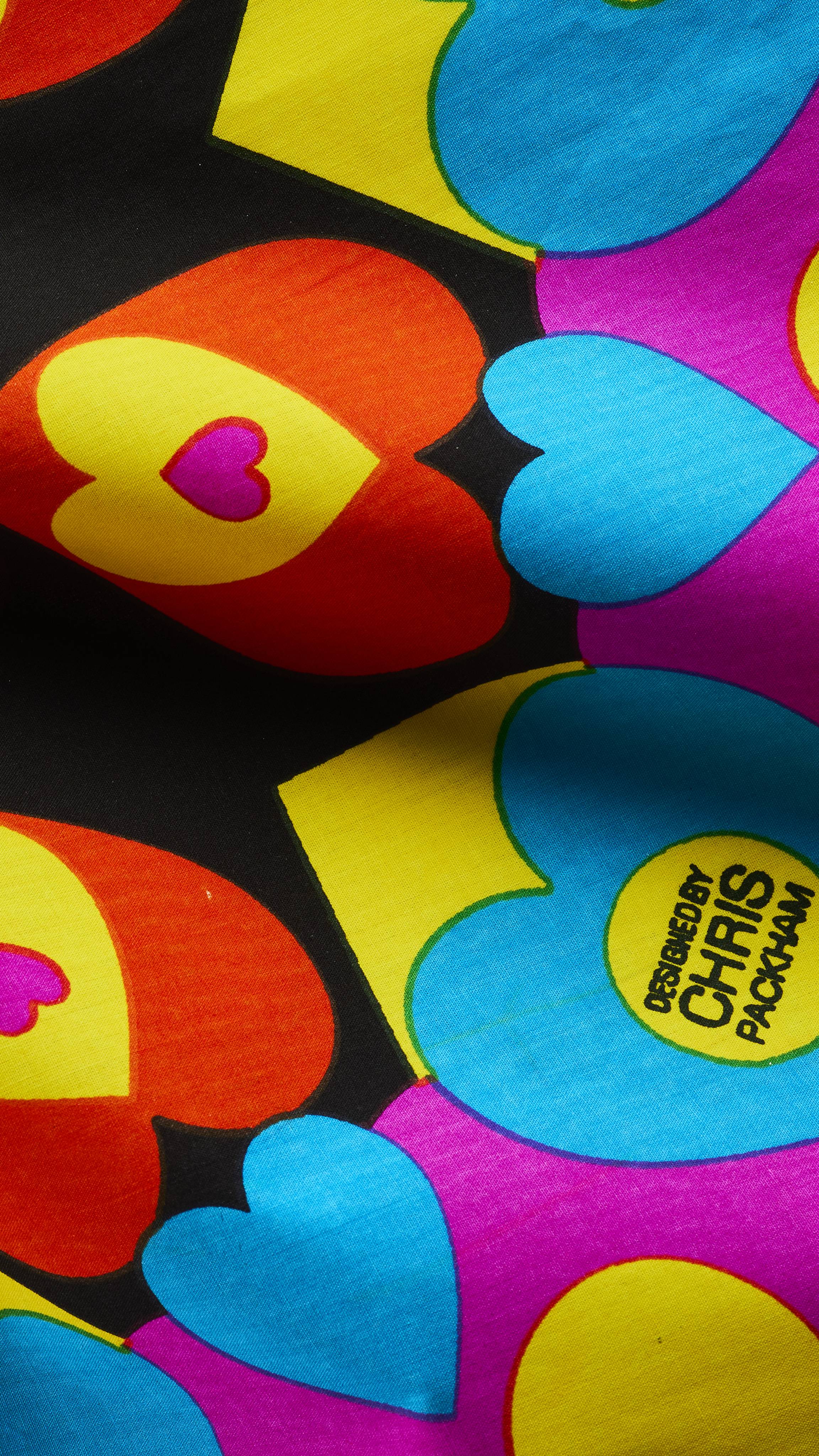A close-up of a vibrant fabric knot wrap with colourful nested hearts in red, yellow, pink, and blue on a black background, featuring a circular detail that reads "DESIGNED BY CHRIS PACKHAM".