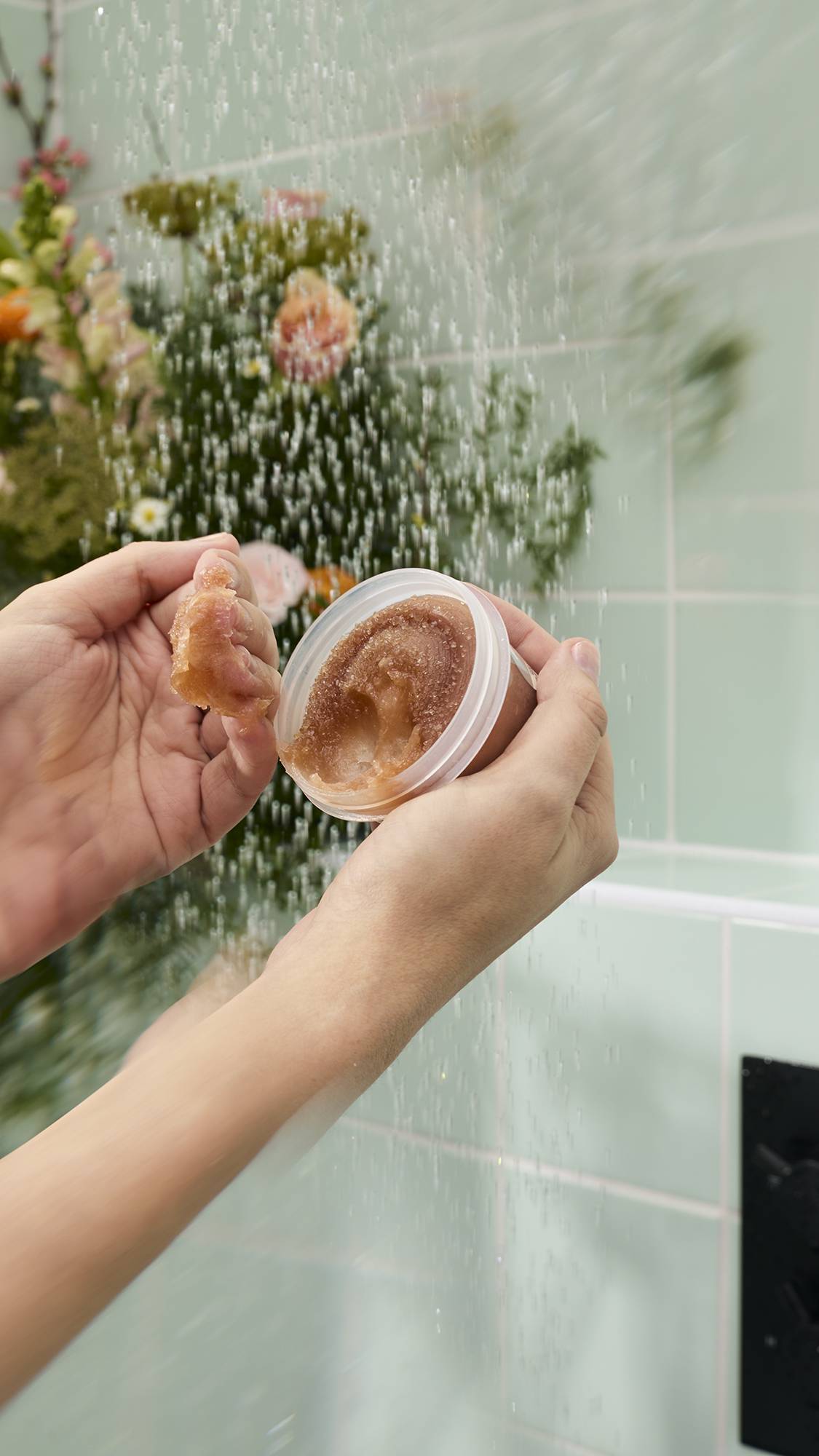 A model's hands hold a small, open pot of Lush "Crème Brûlée" body scrub under a shower. They have scooped out some of the thick, brown, sugary product. The background features pale green tiles and a large, colourful bouquet of flowers.