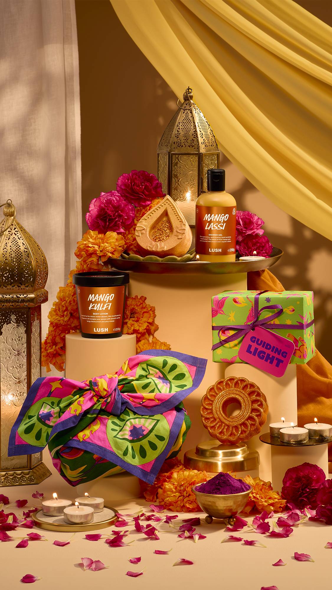 A festive display of Lush products, including Mango Lassi shower gel, Mango Kulfi body lotion, a diya bath bomb, wrapped gifts, and a printed Knot Wrap, set amongst brass lanterns, tealights, and vibrant marigold and rose flowers.