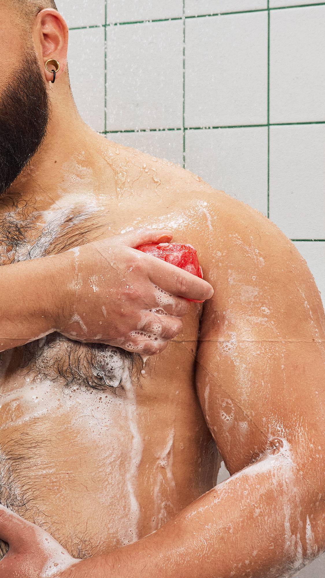 Close-up of a bearded model washing their shoulder with a red soap bar, creating a rich white lather over their chest in a green-grouted tiled shower.