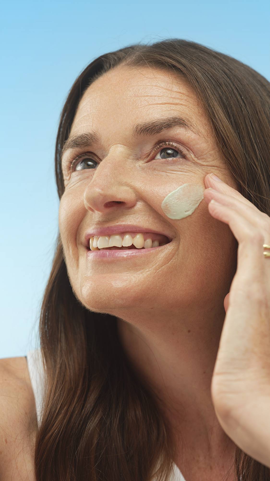 A close-up vertical portrait of a smiling person with long brown hair, looking up while applying a creamy light-green swatch of Feeling Greener primer to their cheek with a finger. The background is a bright blue.