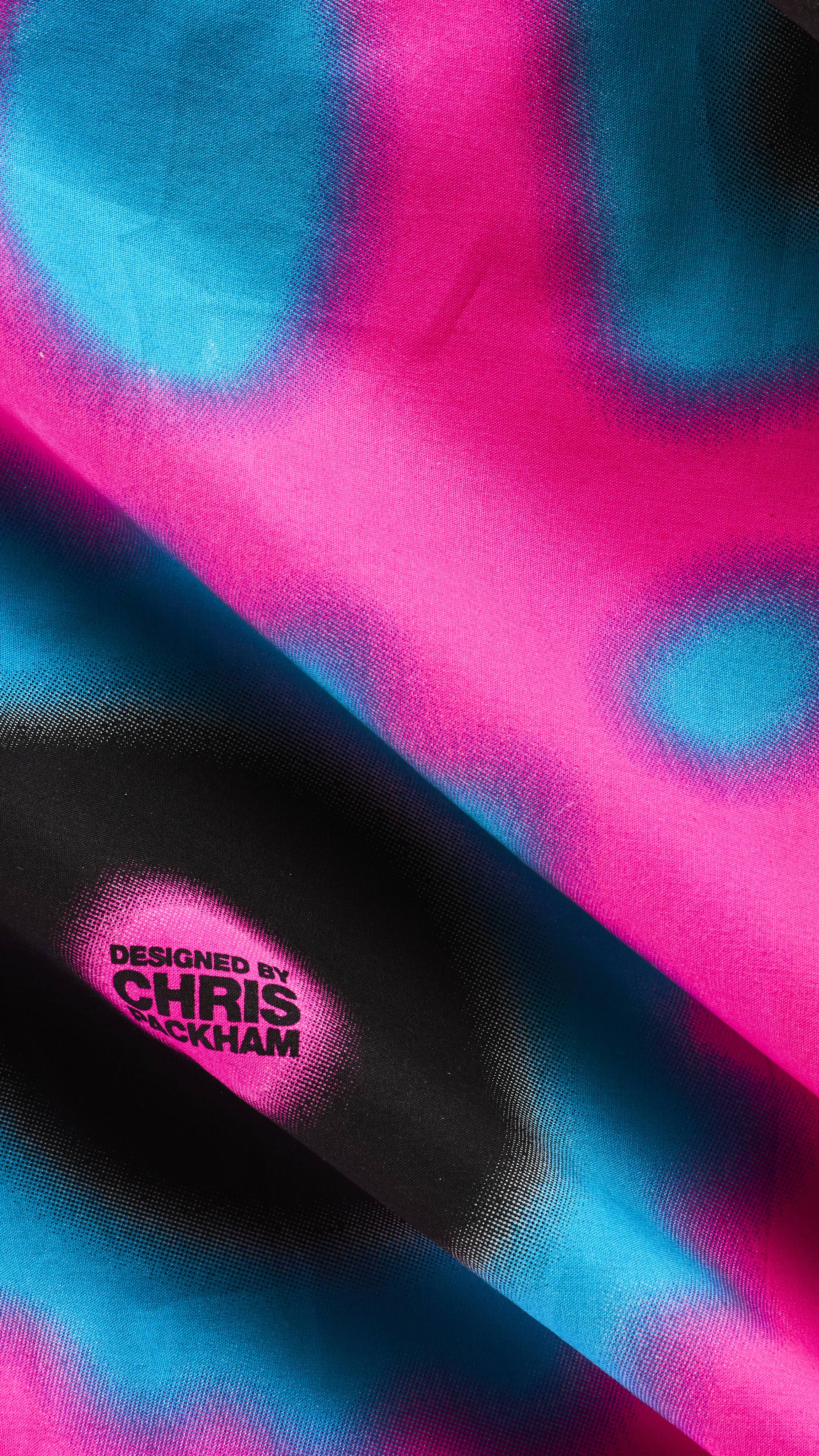 A close-up of a draped fabric Knot Wrap featuring a vibrant, heat-map-style pattern in pink, cyan blue, and black, with black text inside a pink circle reading "DESIGNED BY CHRIS PACKHAM".
